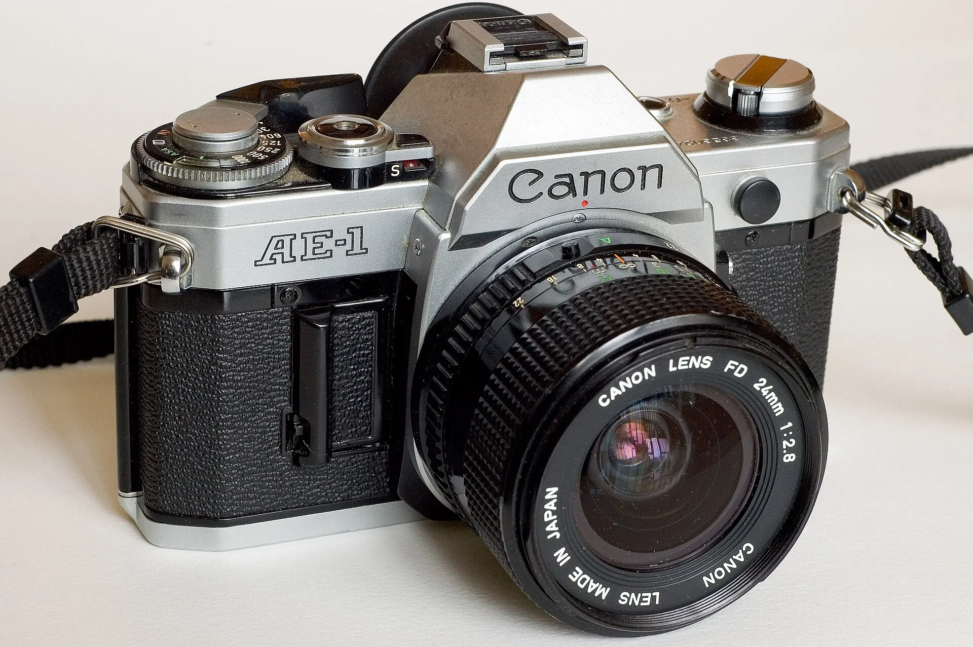 A silver Canon AE-1 35mm film camera with a Canon FD 24mm f/2.8 lens, on a white background