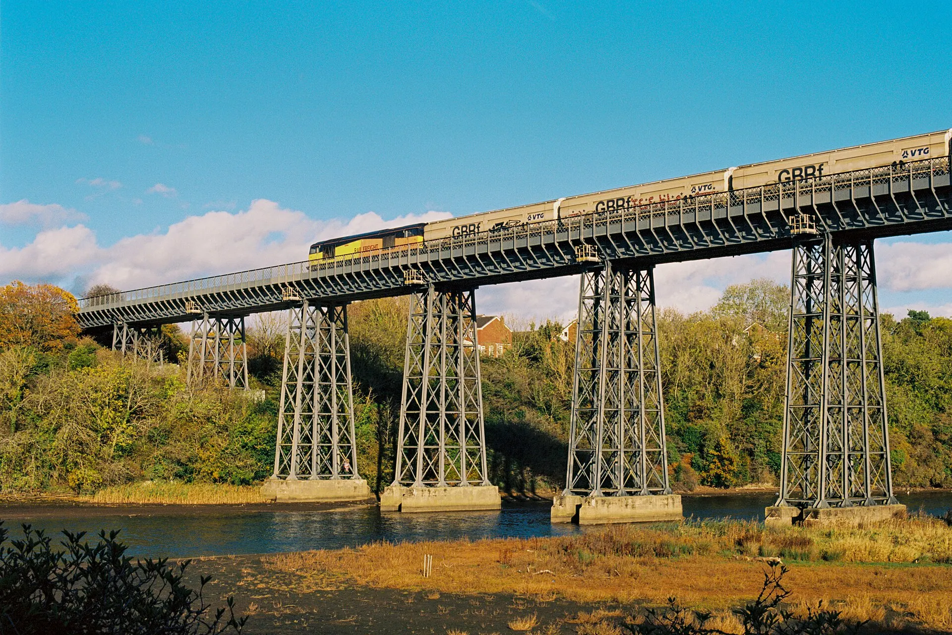 Freight locomotive pulling a train across a tall steel trestle viaduct over a river under a blue sky