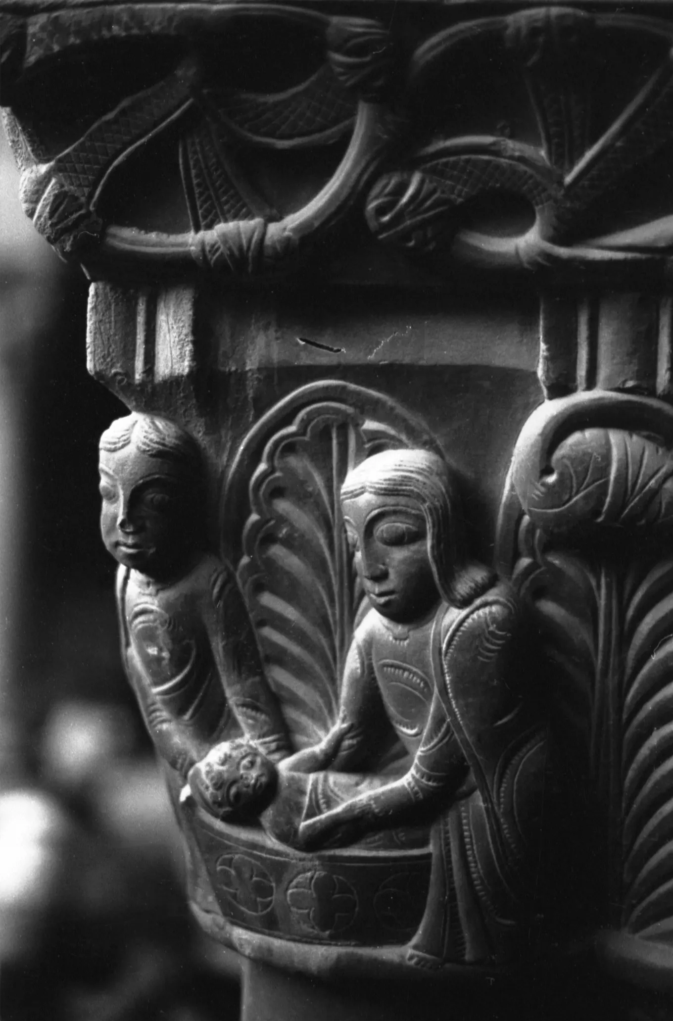 Black-and-white close-up of a carved stone column capital with figures and leaves