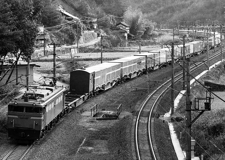 Black and white view of a freight train on a curving mountain line with a pusher locomotive at the rear