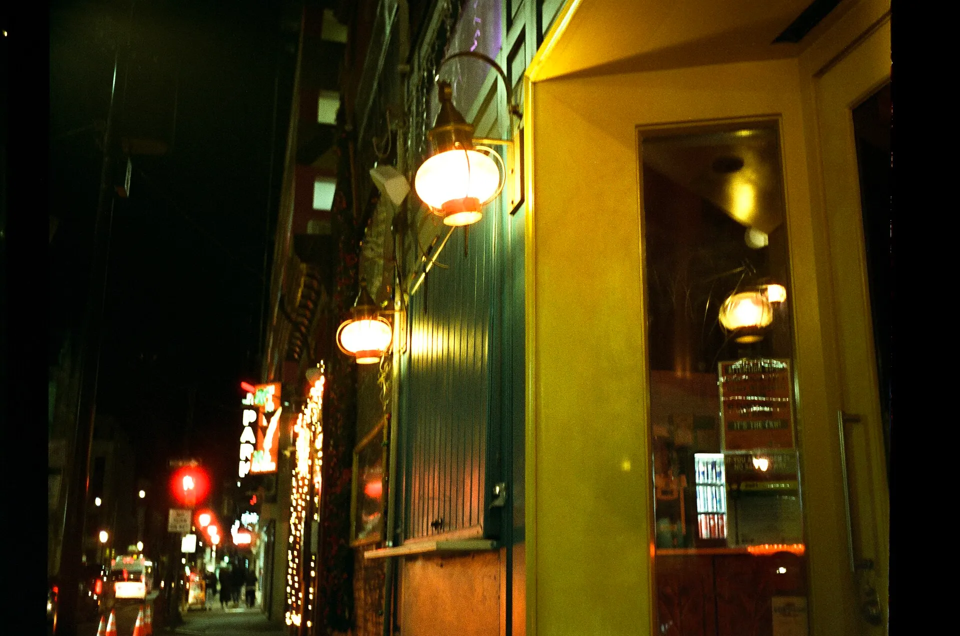 A narrow city street at night with glowing wall lanterns, a lit shop window and red lights in the distance