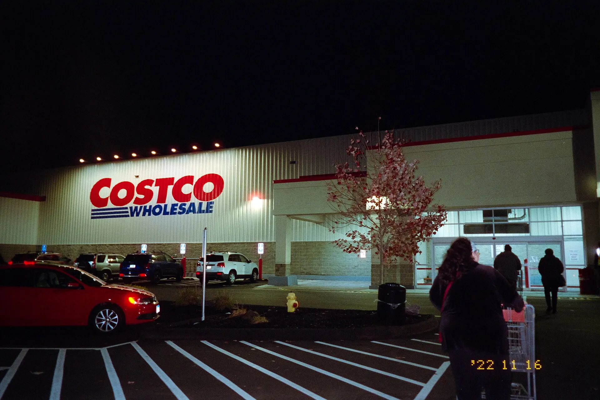 A Costco Wholesale store lit up at night, with a red car and shoppers with carts in the parking lot
