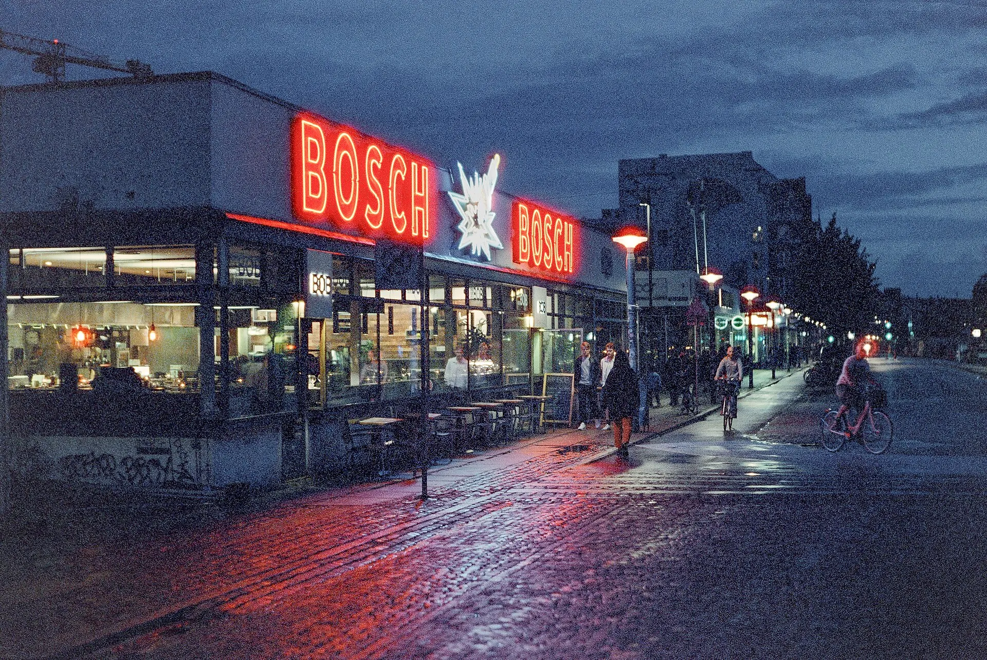 The Bosch building in Copenhagen's Kødbyen at dusk, red neon letters glowing over a wet street with cyclists