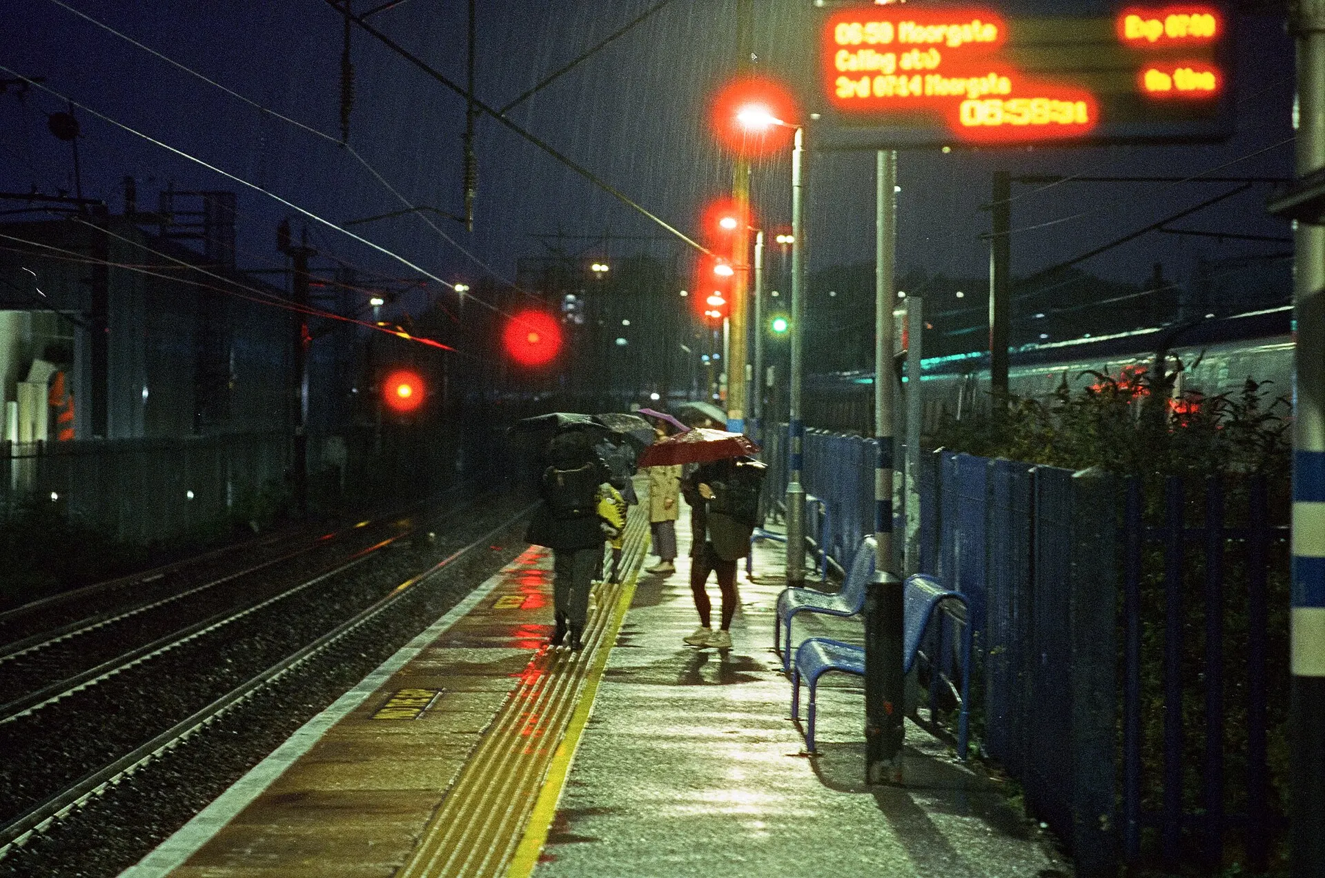 A rain-wet platform at Hornsey railway station in the dark, red signal lights and orange departure boards glowing