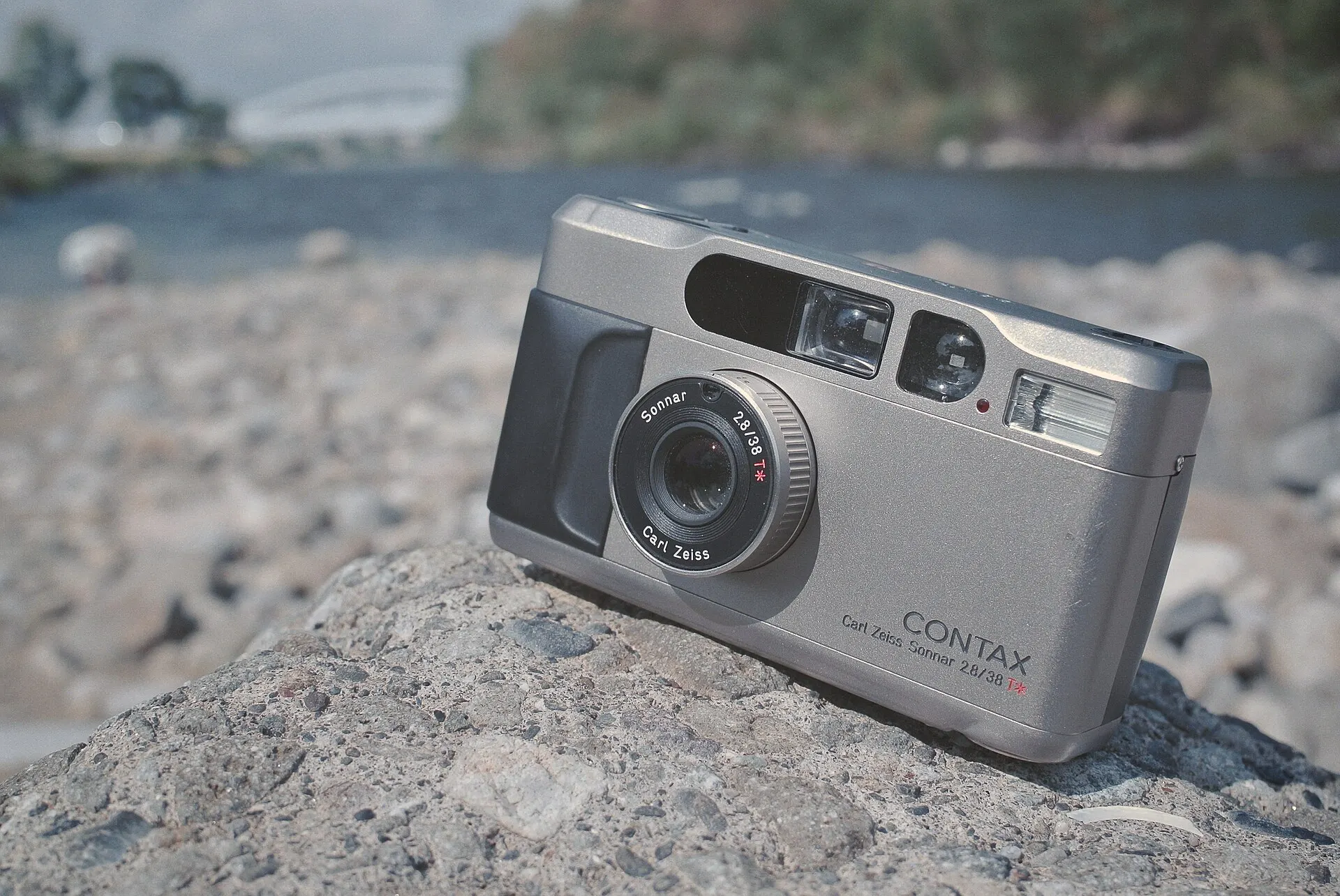 A champagne silver Contax T2 with its lens extended, resting on a rock by a river