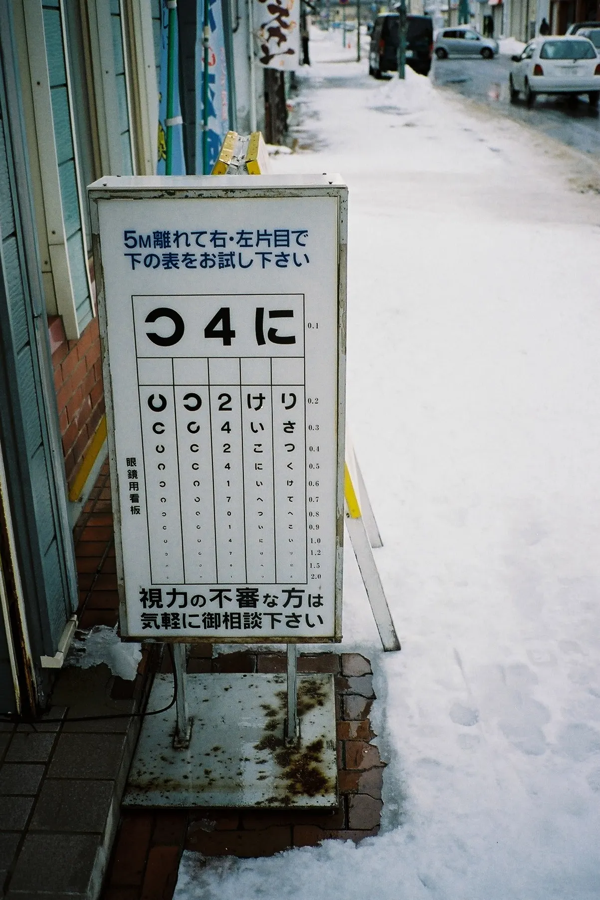 An eye-test chart sign standing in snow outside an old shopping street