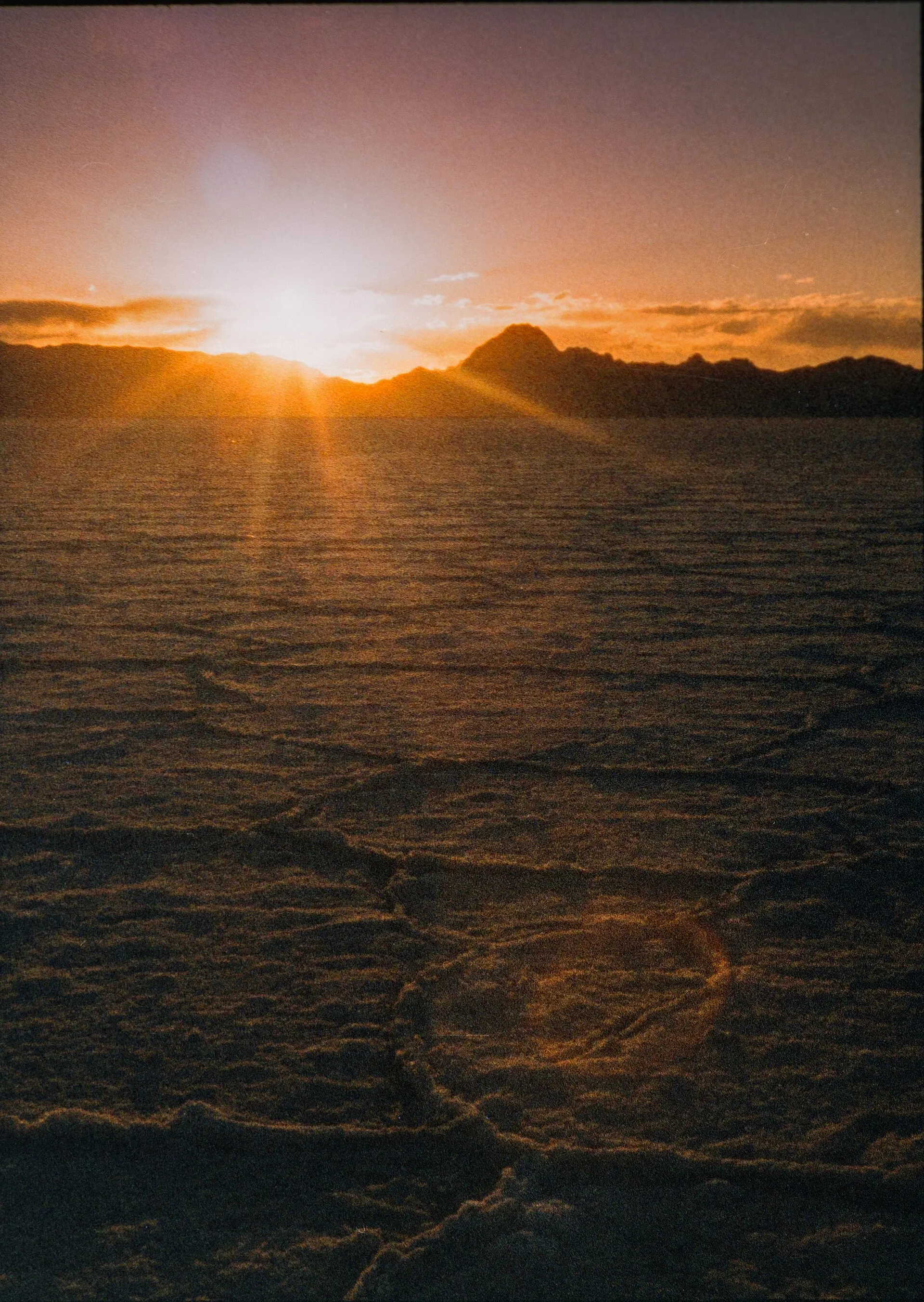 The sun setting behind mountains over cracked salt flats, with a strong orange glow around the sun