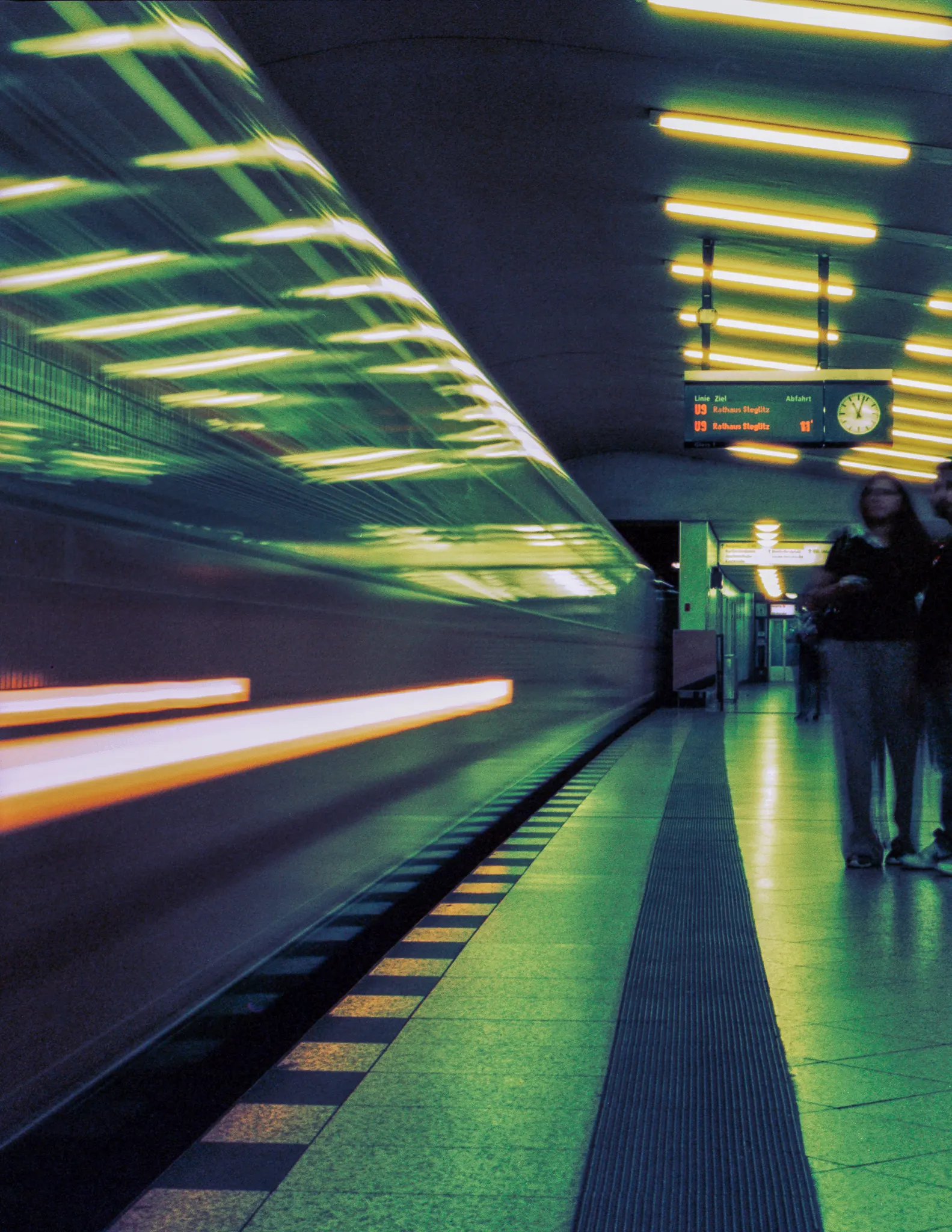 A subway train streaking past a platform under green-lit ceiling lights in a long exposure