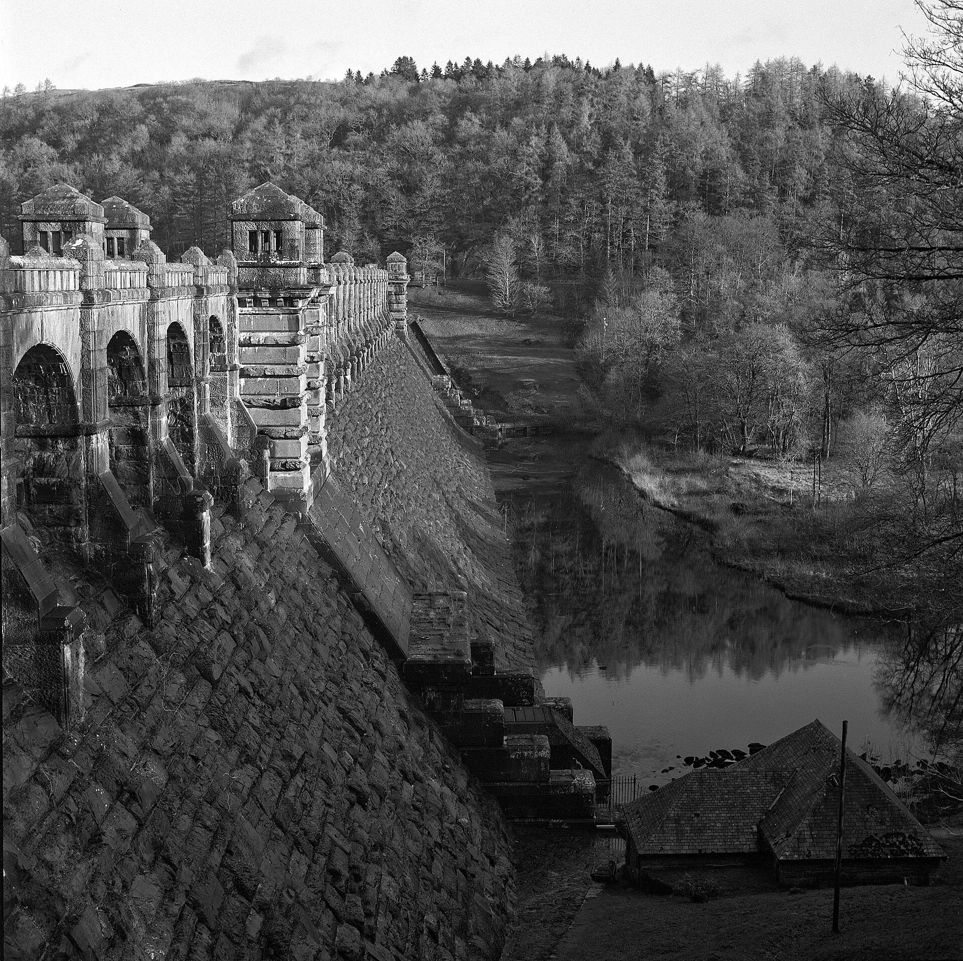 Black and white view down the face of the Lake Vyrnwy dam, with its arched stone towers, the water and wooded hills behind