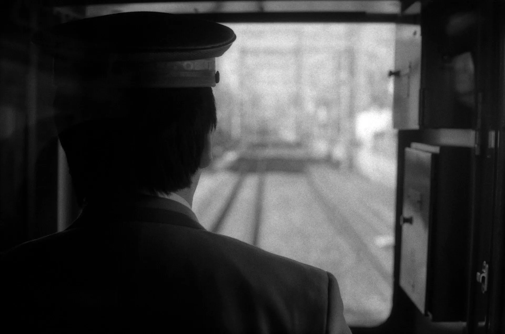 A train conductor in a cap seen from behind, looking out of the cab window down the track