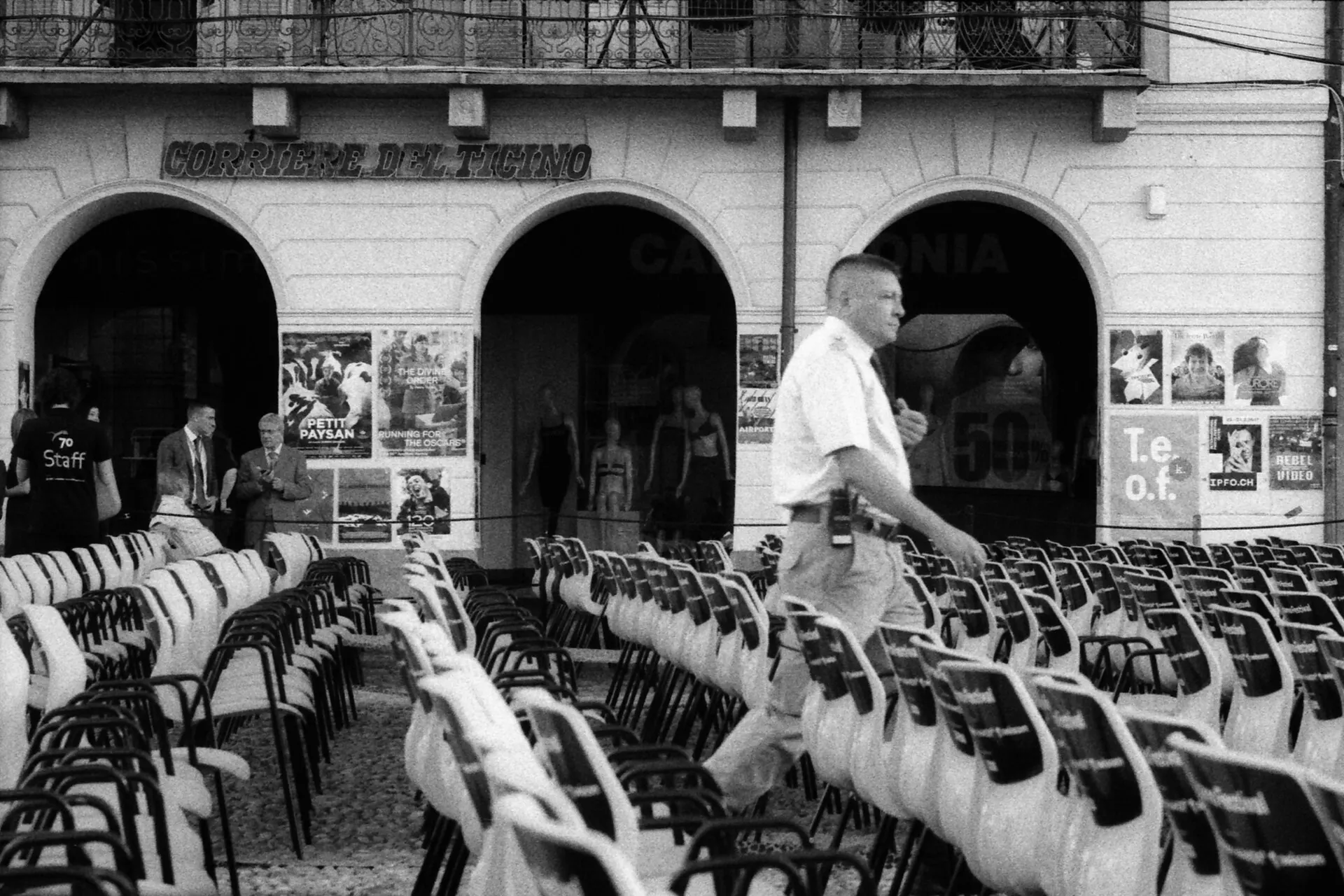 Rows of white chairs in Piazza Grande in daylight, with a man walking past the arcades behind them