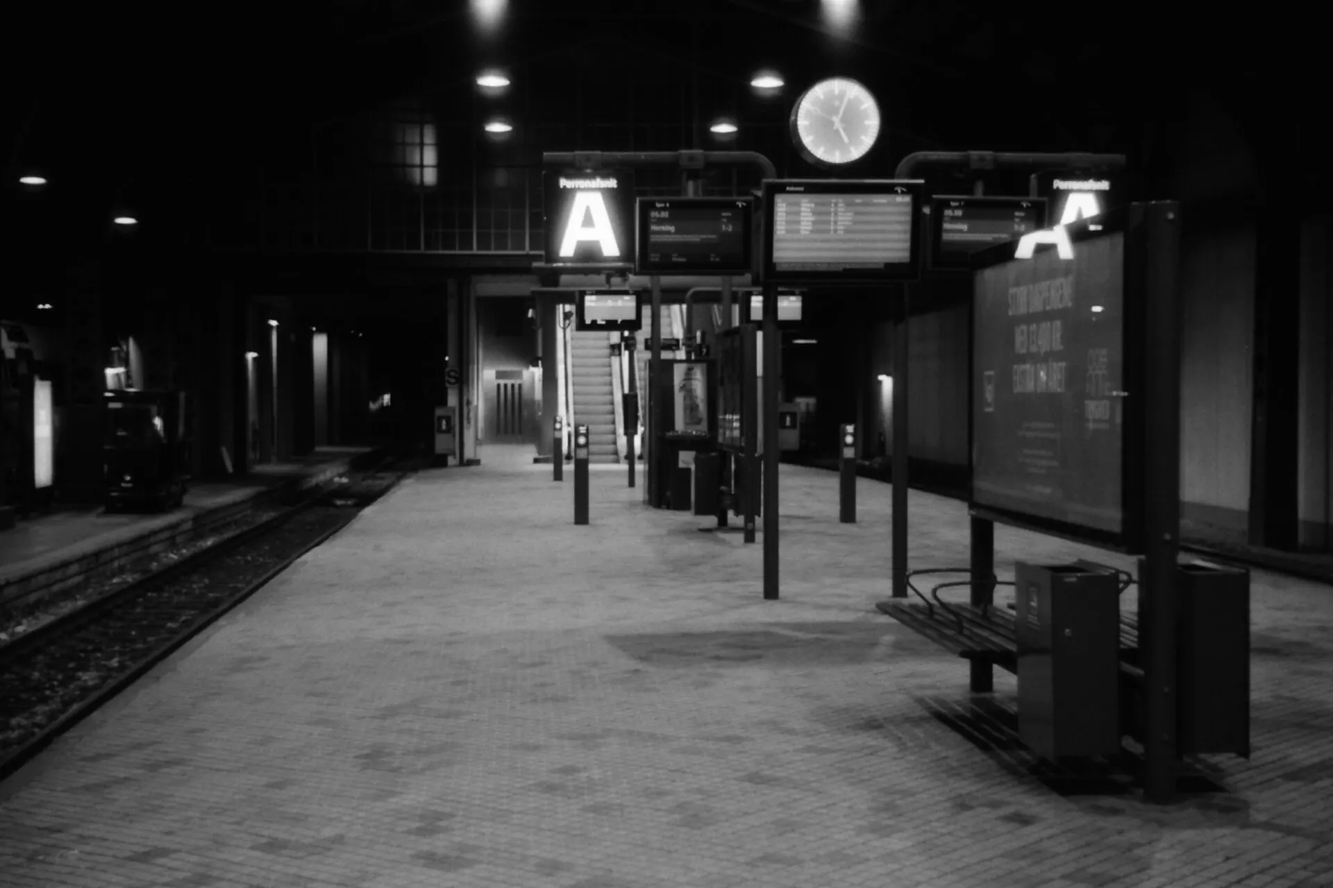 Black and white photo of an empty train platform at night under overhead lights, with a clock and station signs