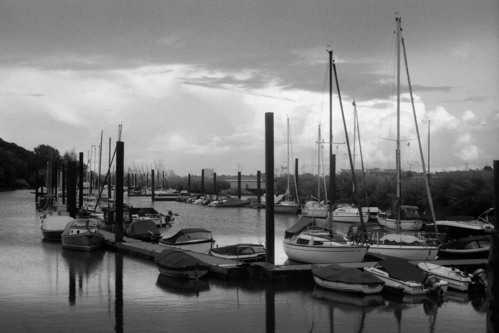 Black and white photo of sailboats moored in a small harbor under a cloudy sky