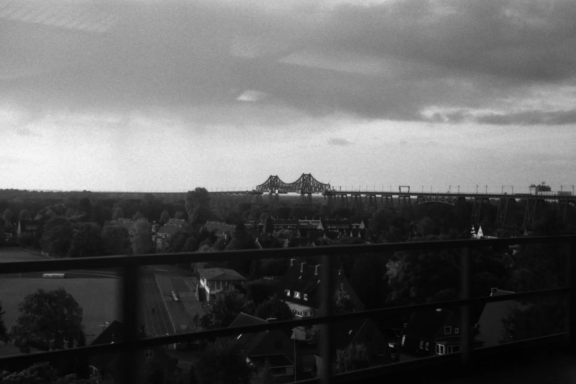 Black and white photo of a long steel railway bridge on the horizon, seen over trees from a moving train