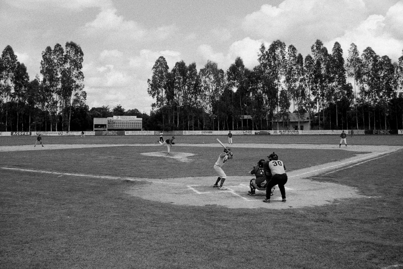 Black and white photo of a baseball game seen from behind home plate, with a batter, catcher and umpire and tall trees behind the field