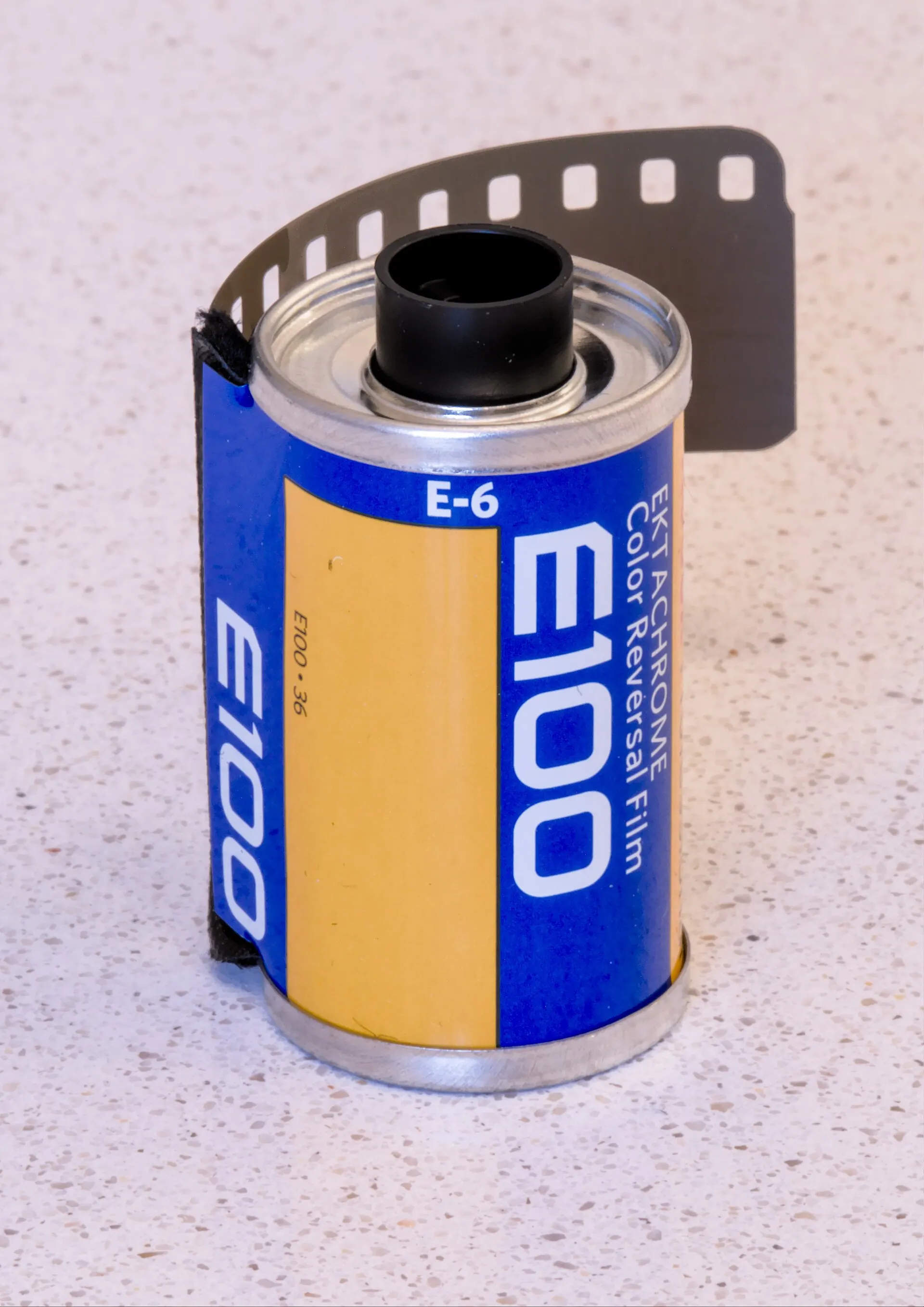 A 35mm cartridge of Kodak Ektachrome E100 slide film, E-6, with the film leader pulled out, on a plain light background