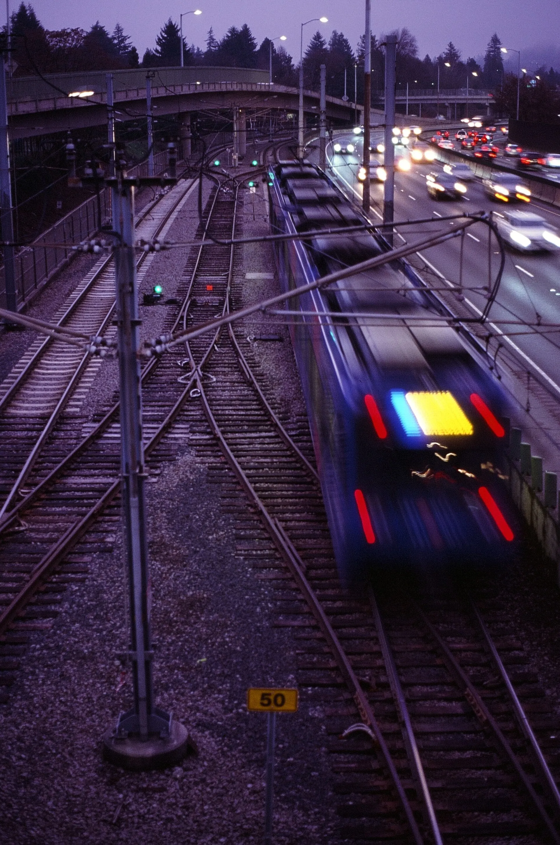 A TriMet MAX light rail train blurred as it leaves a station beside a freeway at dusk