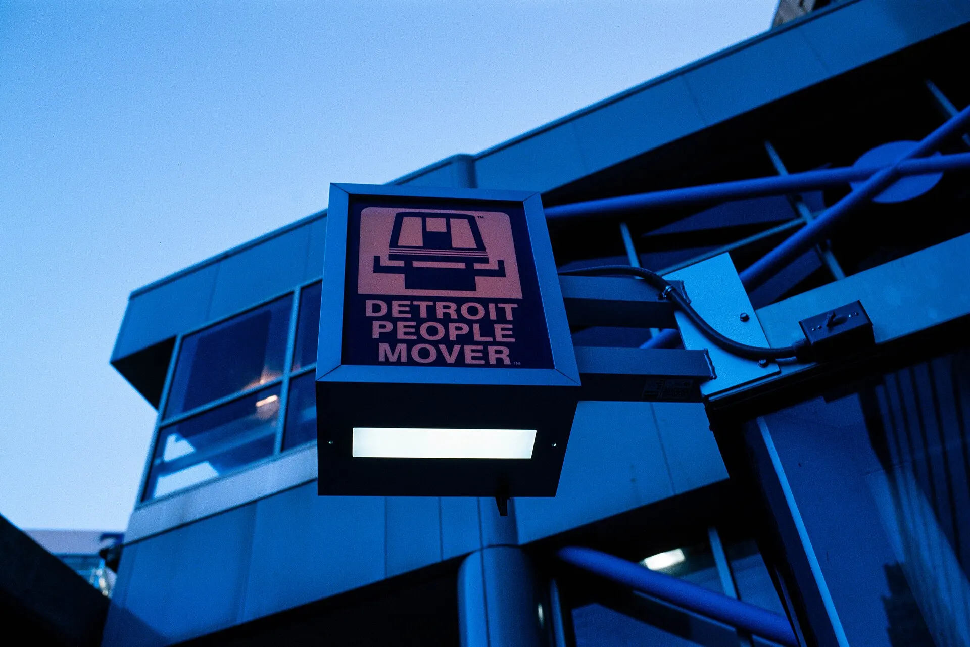 A Detroit People Mover station sign on a blue steel structure against a deep blue sky