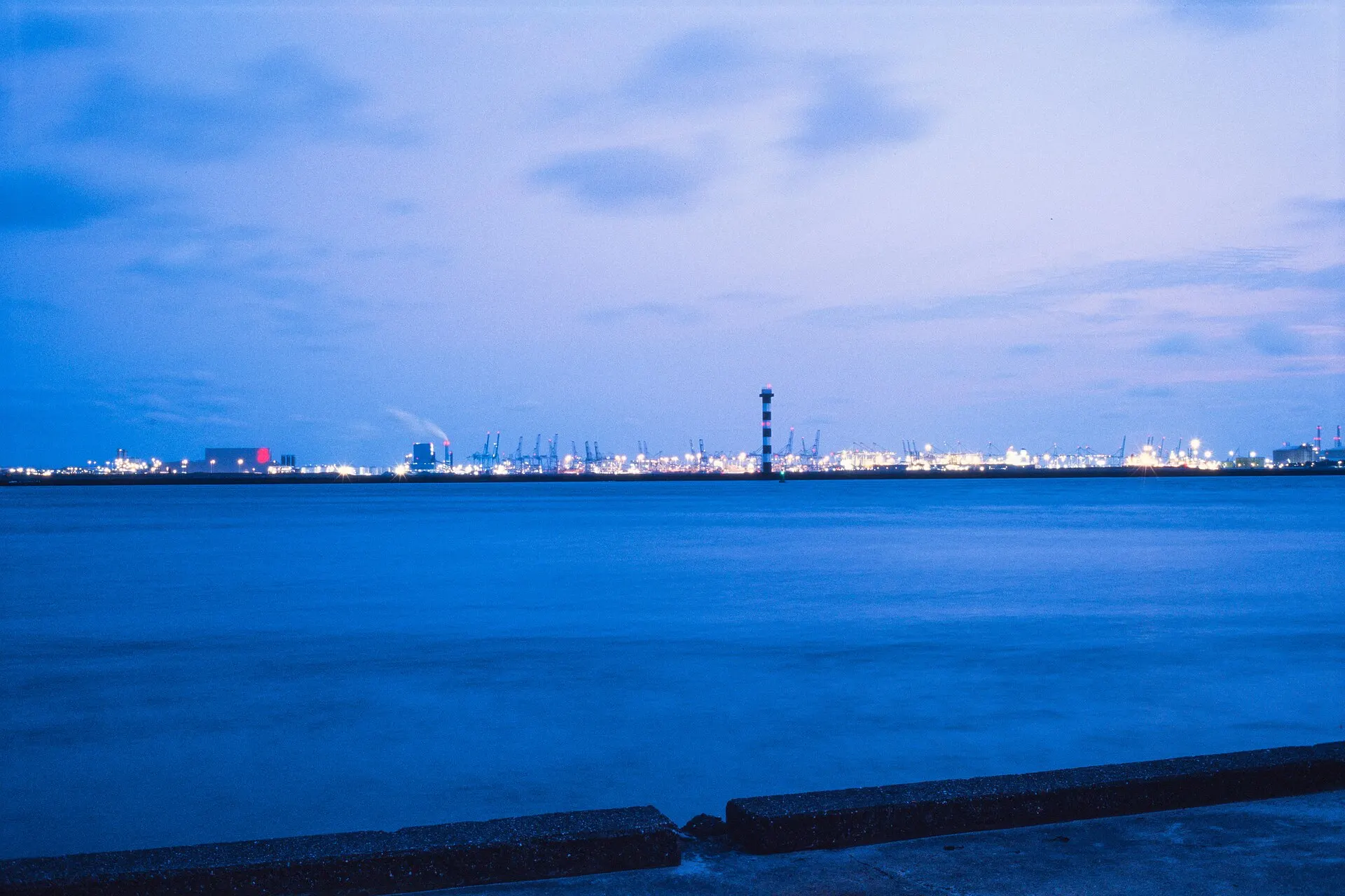 A calm sea at blue hour with a distant industrial shoreline lit on the horizon