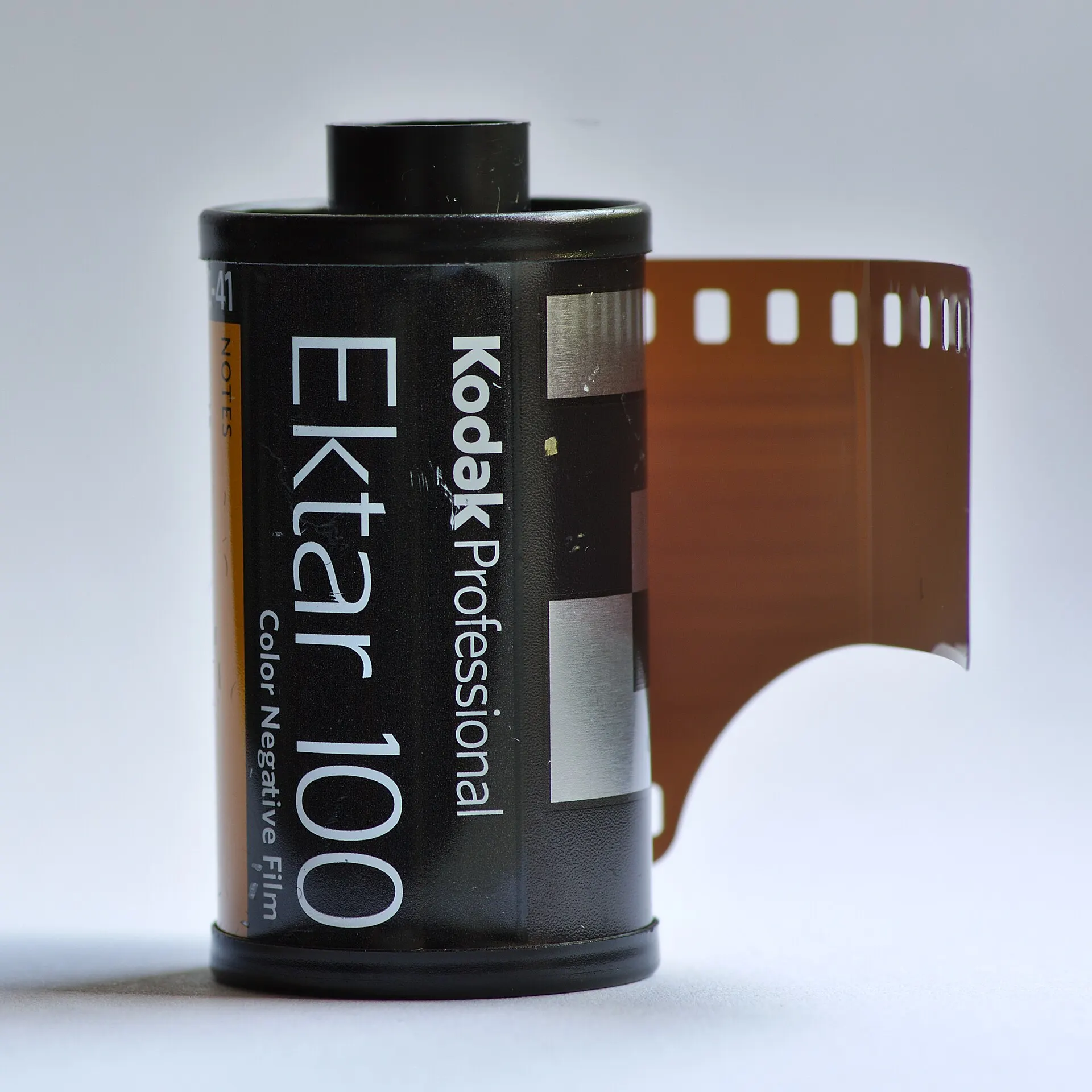 A 35mm cartridge of Kodak Professional Ektar 100 color negative film with the leader pulled out, on a plain light background