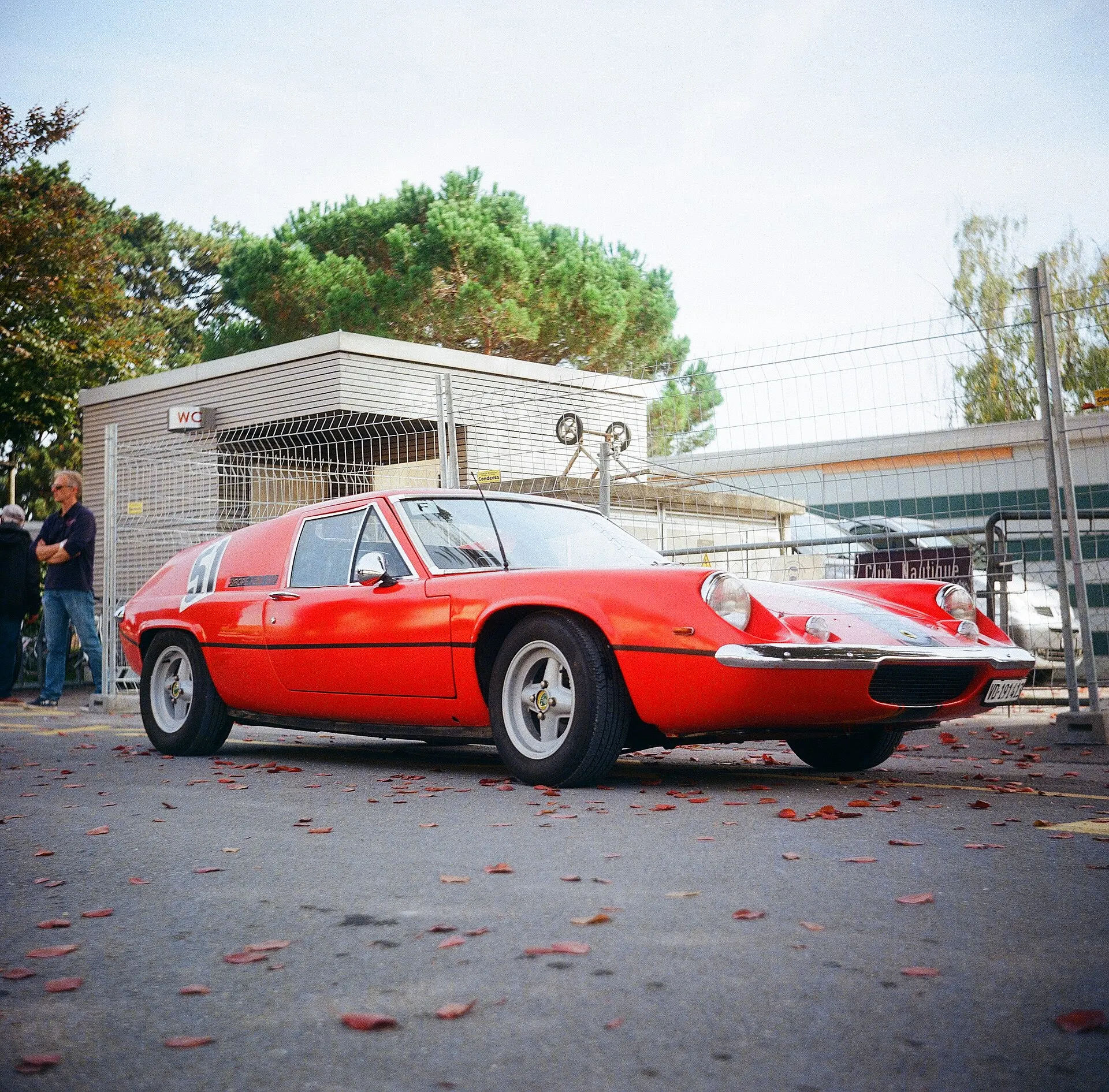 A red Lotus Europa sports car parked on a street lined with fallen red leaves