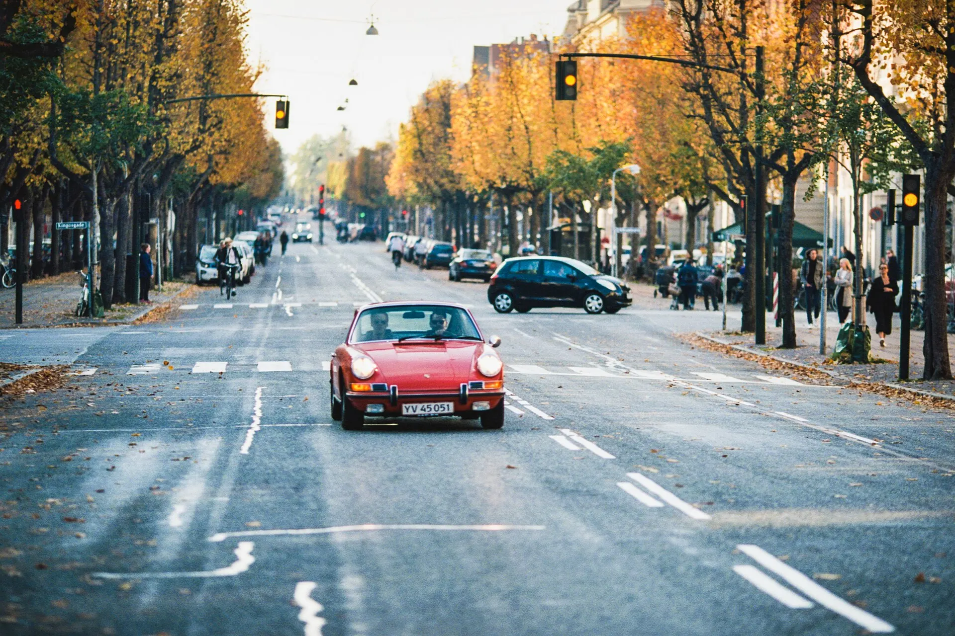 A red vintage Porsche 911 driving down a tree-lined avenue with yellow autumn leaves