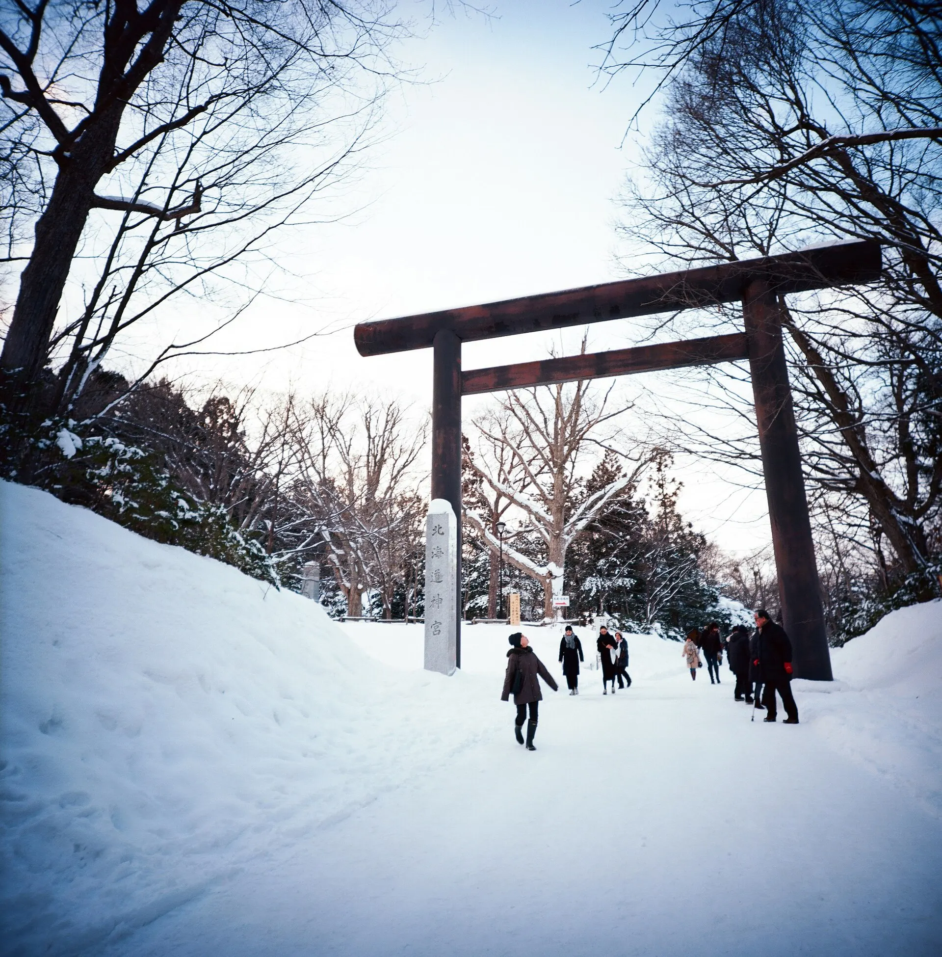 A wooden torii gate over a snowy path with tall trees and a few people walking