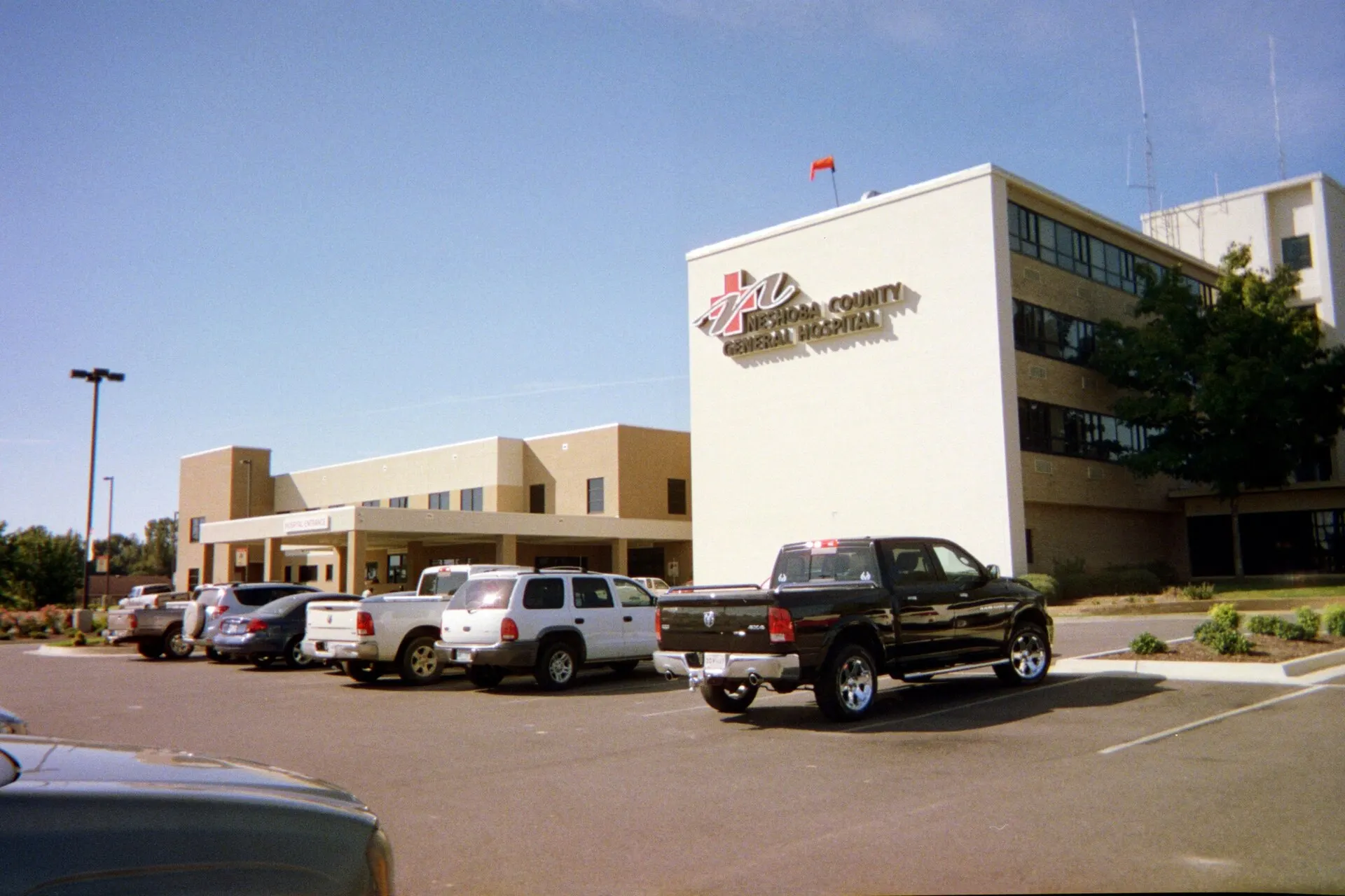 A white hospital building with a red sign and pickup trucks in the parking lot under a clear blue sky