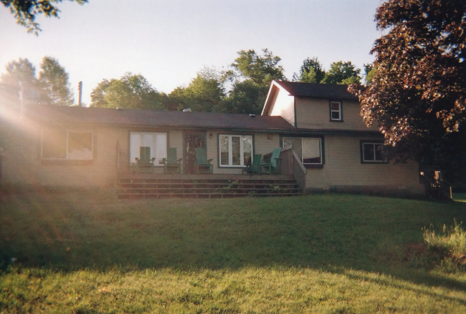 A two-story lake house with a wooden deck and lawn, with sun flare coming in from the left