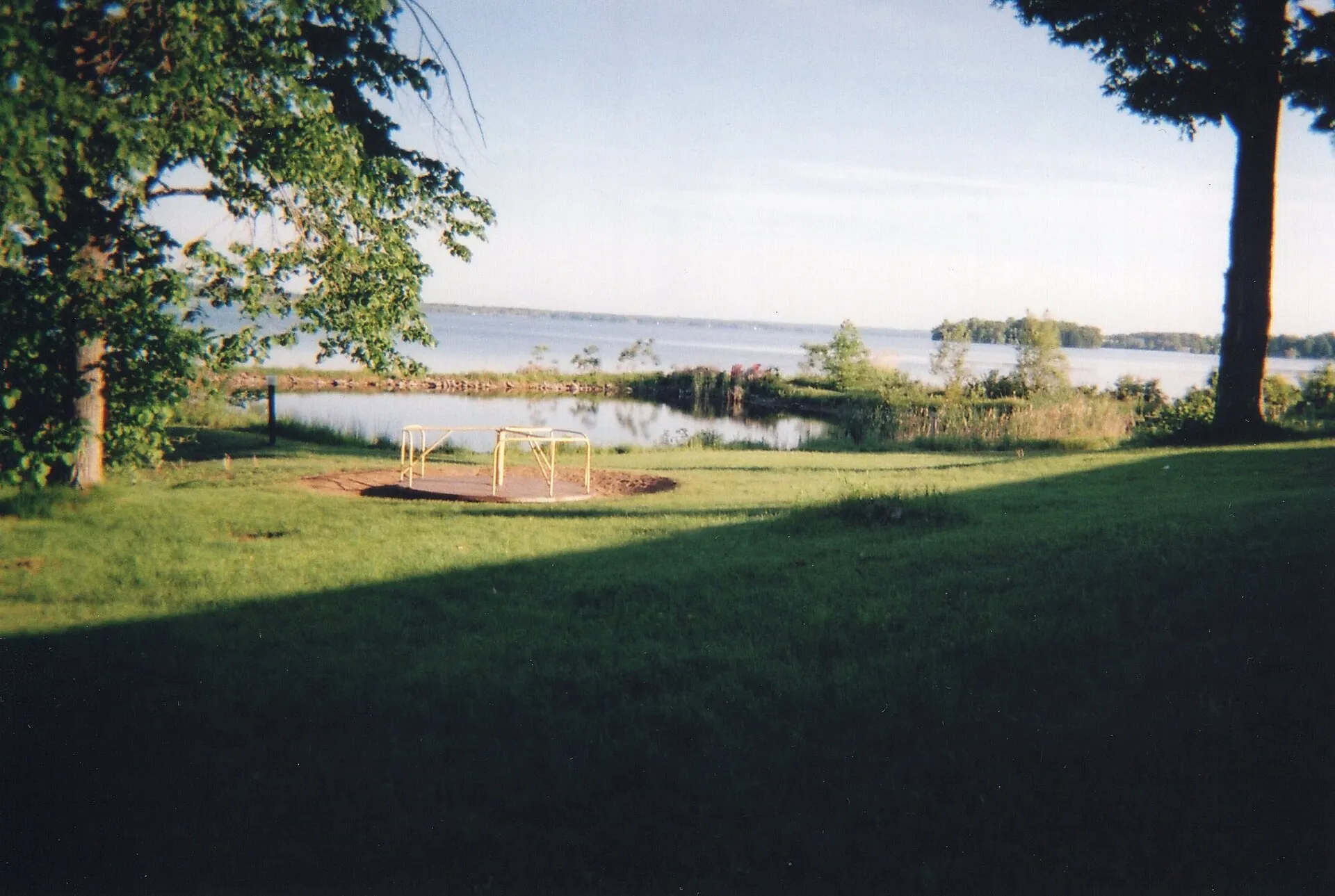A shaded lawn in the foreground leading to a sunlit lake framed by trees
