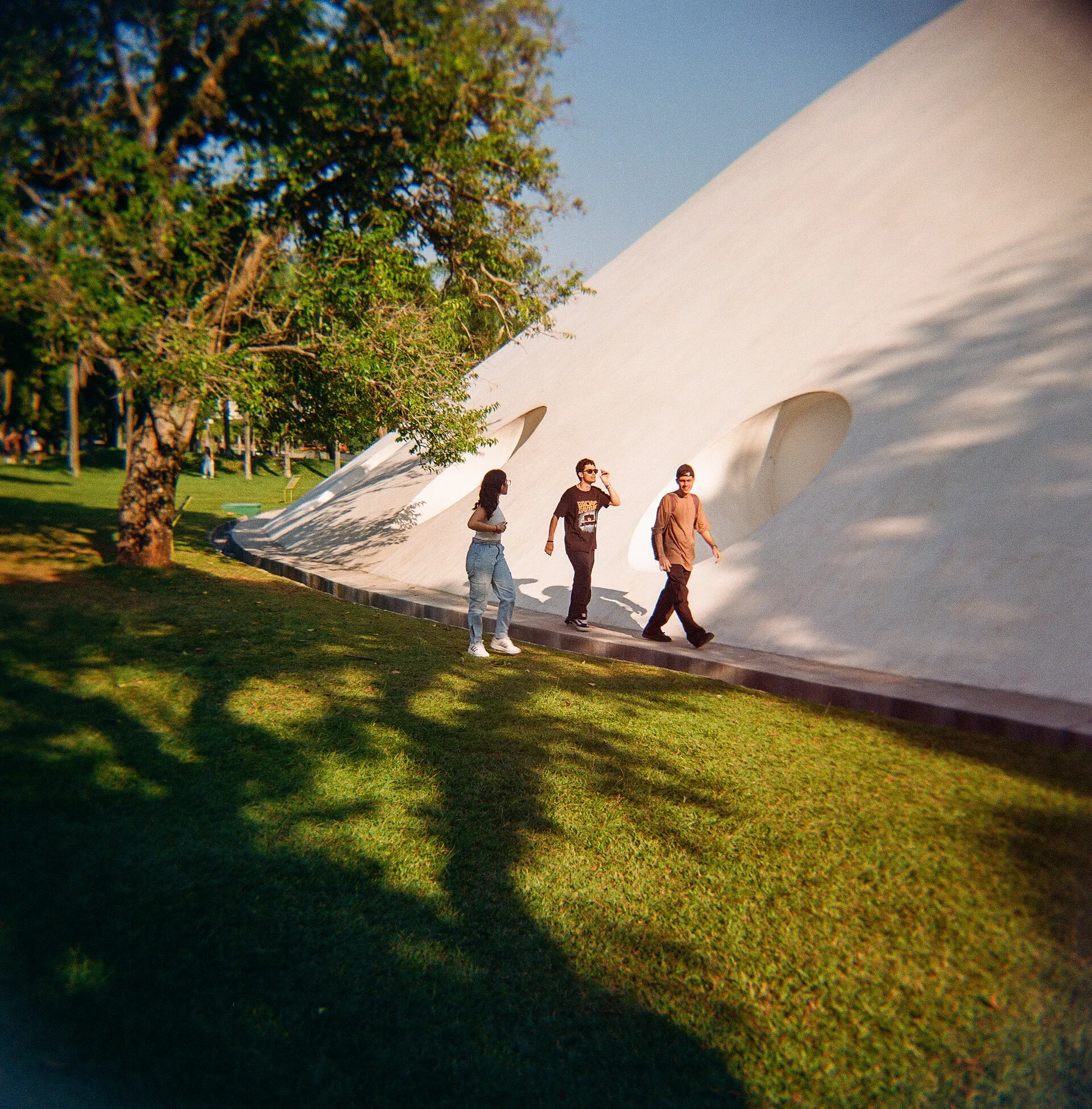 White curved dome of the Oca pavilion beside trees, with three people walking on sunlit grass