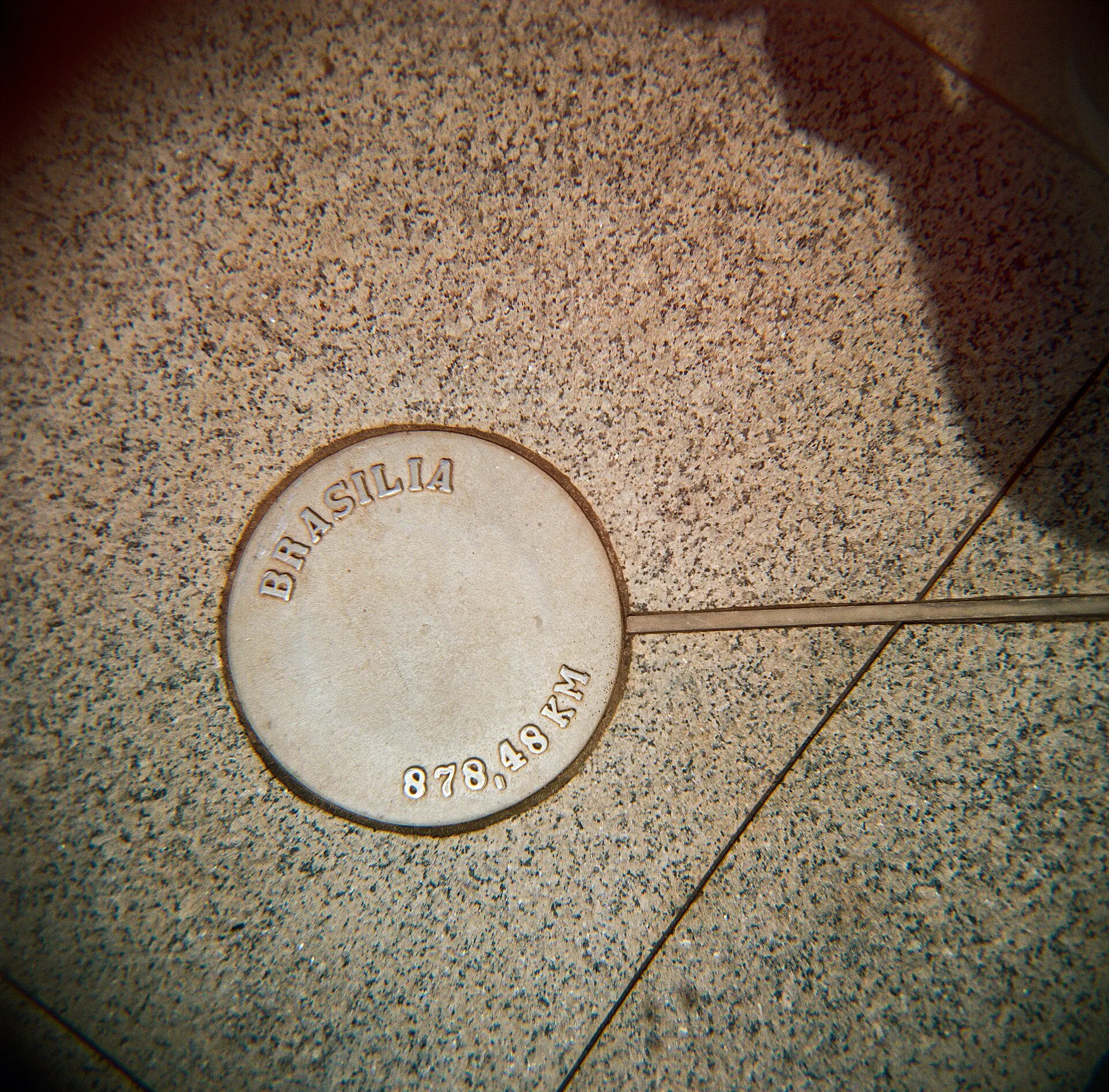 Round metal marker inscribed Brasilia set into speckled stone pavement, with dark corners from a Holga lens