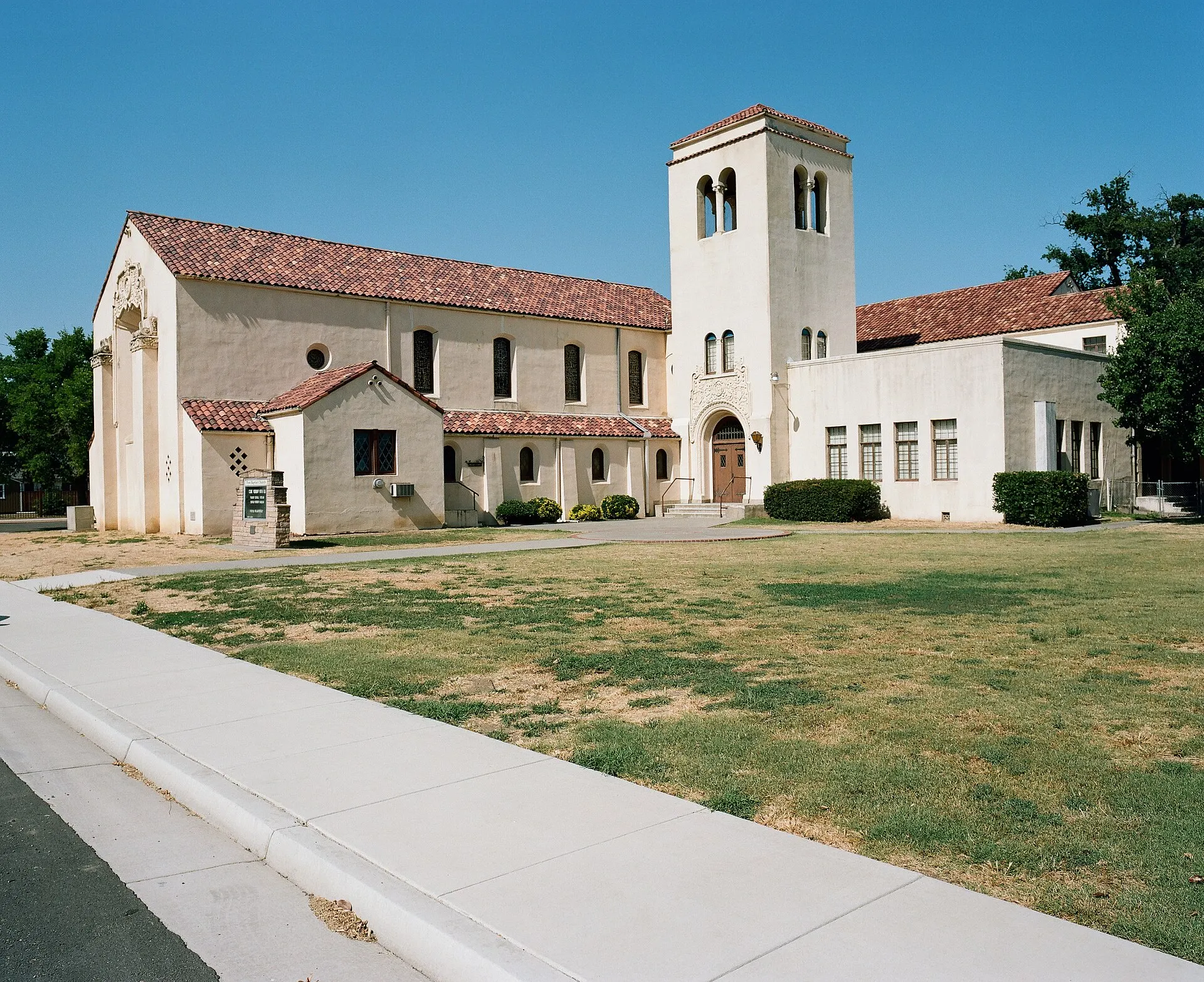 Cream stucco church with a bell tower and red tile roof under a clear blue sky, with a lawn and sidewalk in front