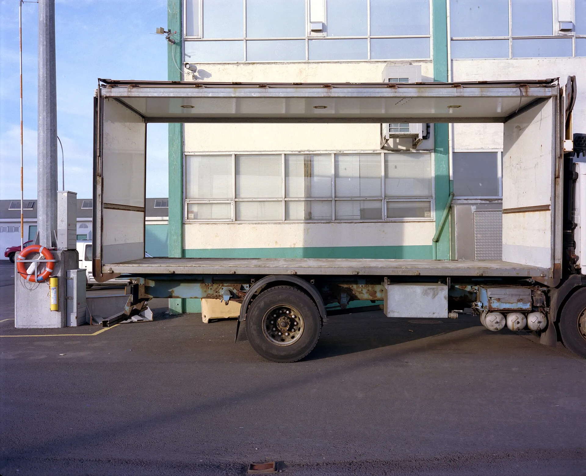 A box truck with open sides parked on a street in front of a glass-fronted building