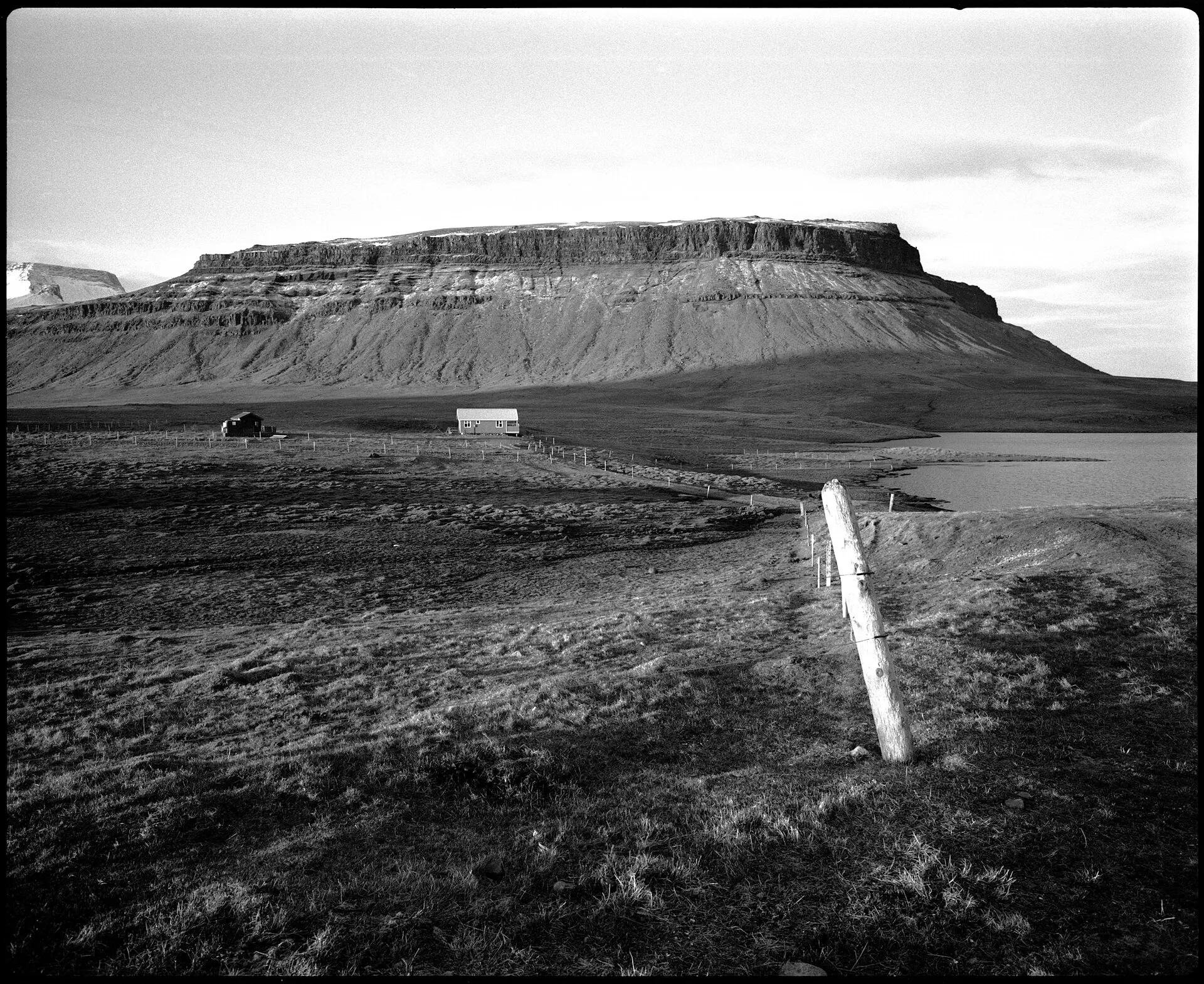 Flat-topped mountain above open grassland and a fence post under a pale sky, in high-contrast black and white