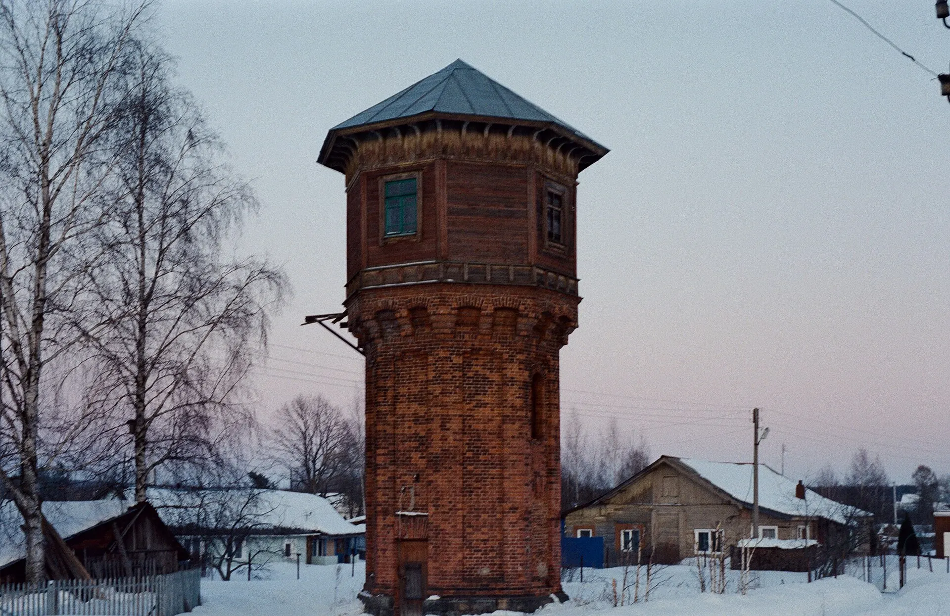 Red-brick railway water tower beside snowy houses and birch trees at dusk, shot on Kodak Vision3 250D