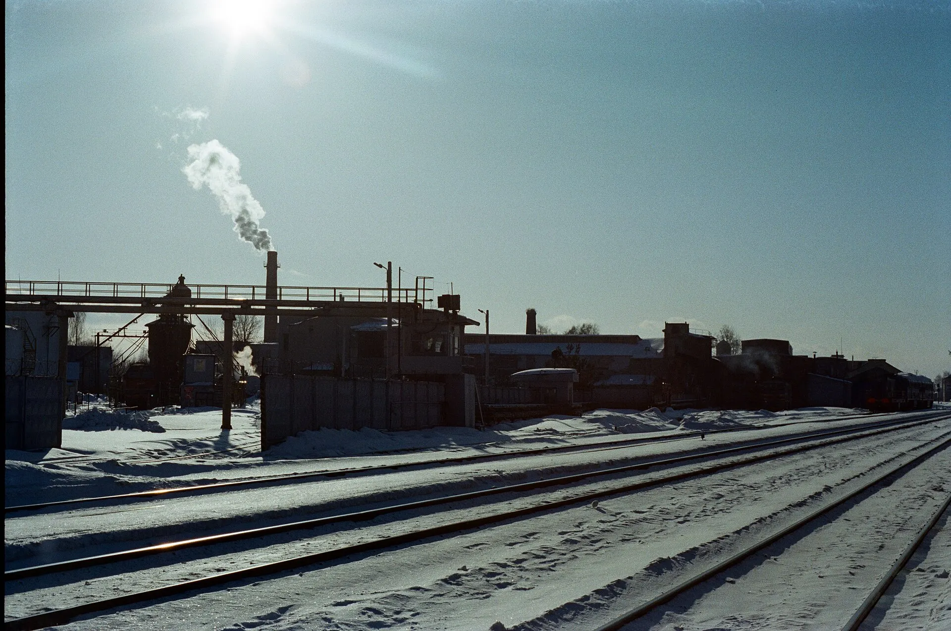 Snow-covered railway tracks and an industrial building with a smoking chimney under bright sun
