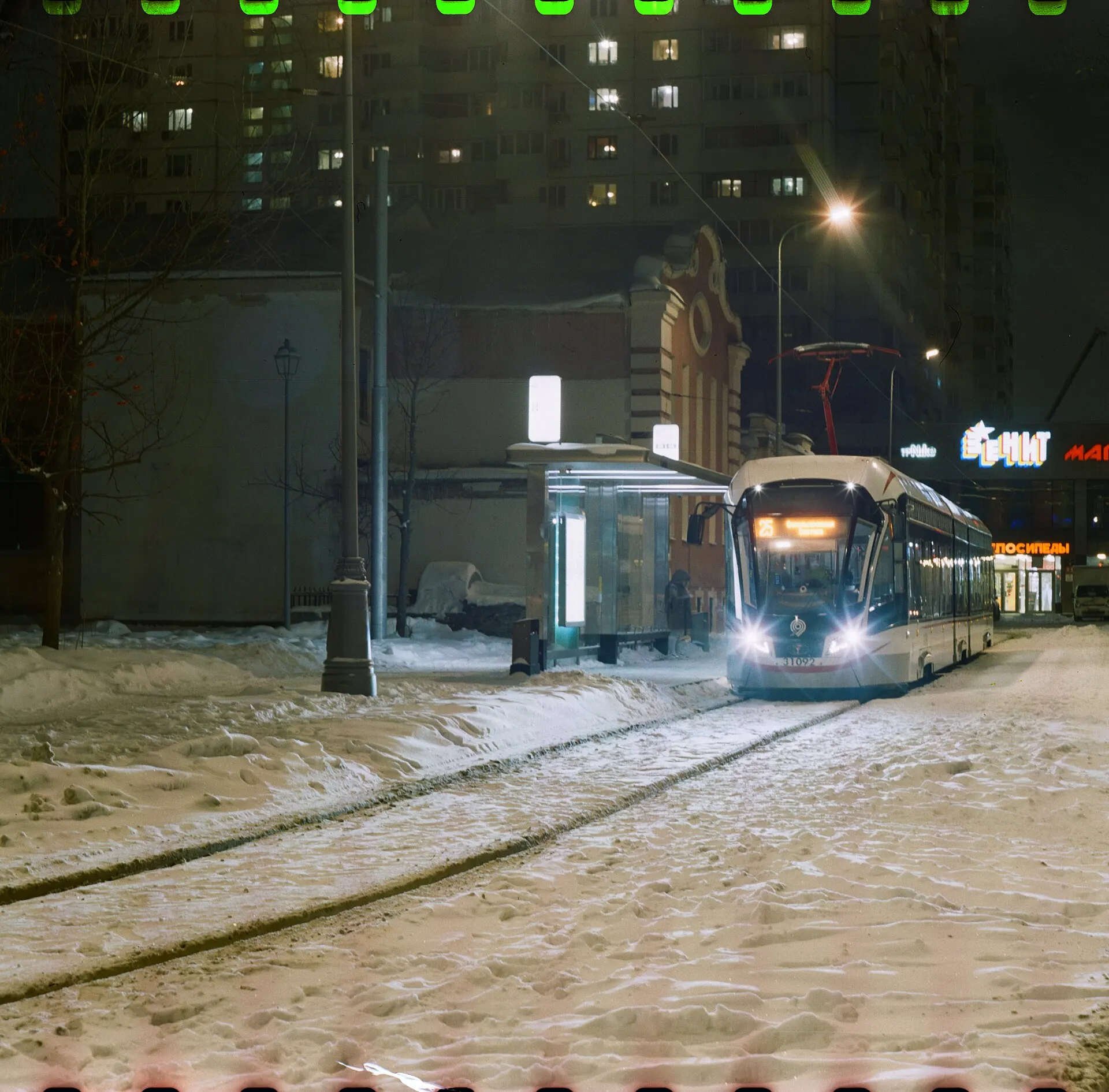 A modern tram stopped at a lit shelter on a snowy square at night