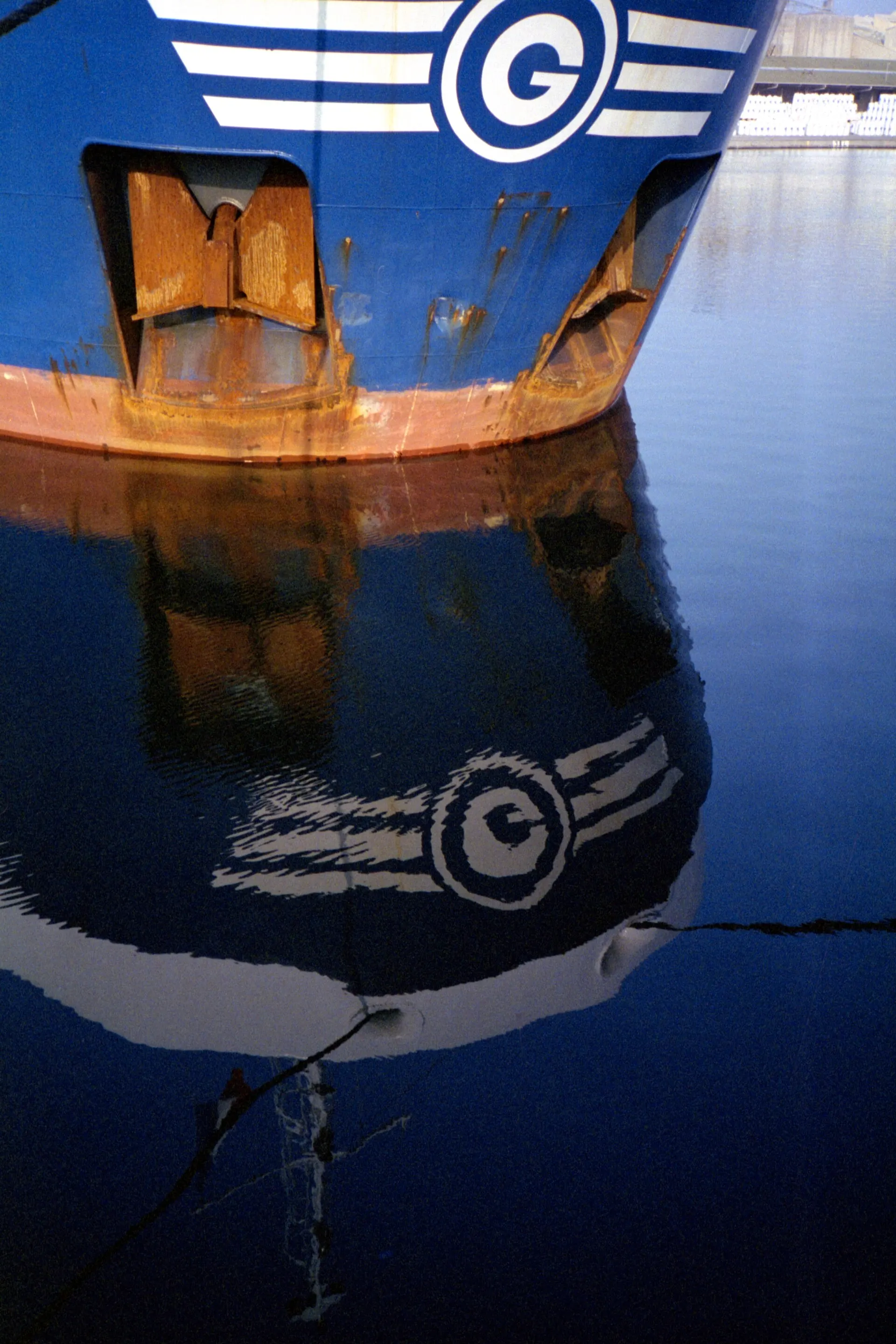 Blue ship hull with a rusty bow and a white G logo reflected in calm harbor water