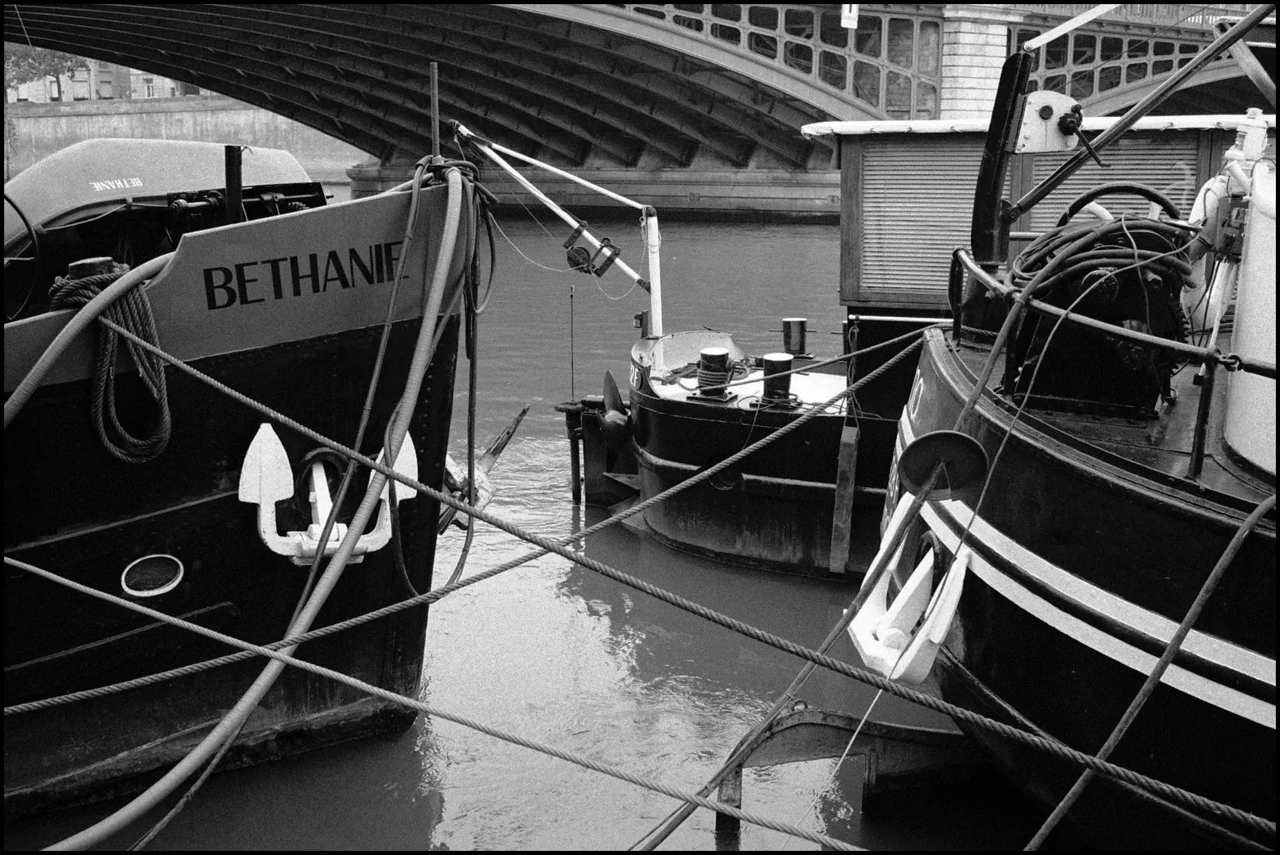 Black and white photo of moored river barges under a bridge