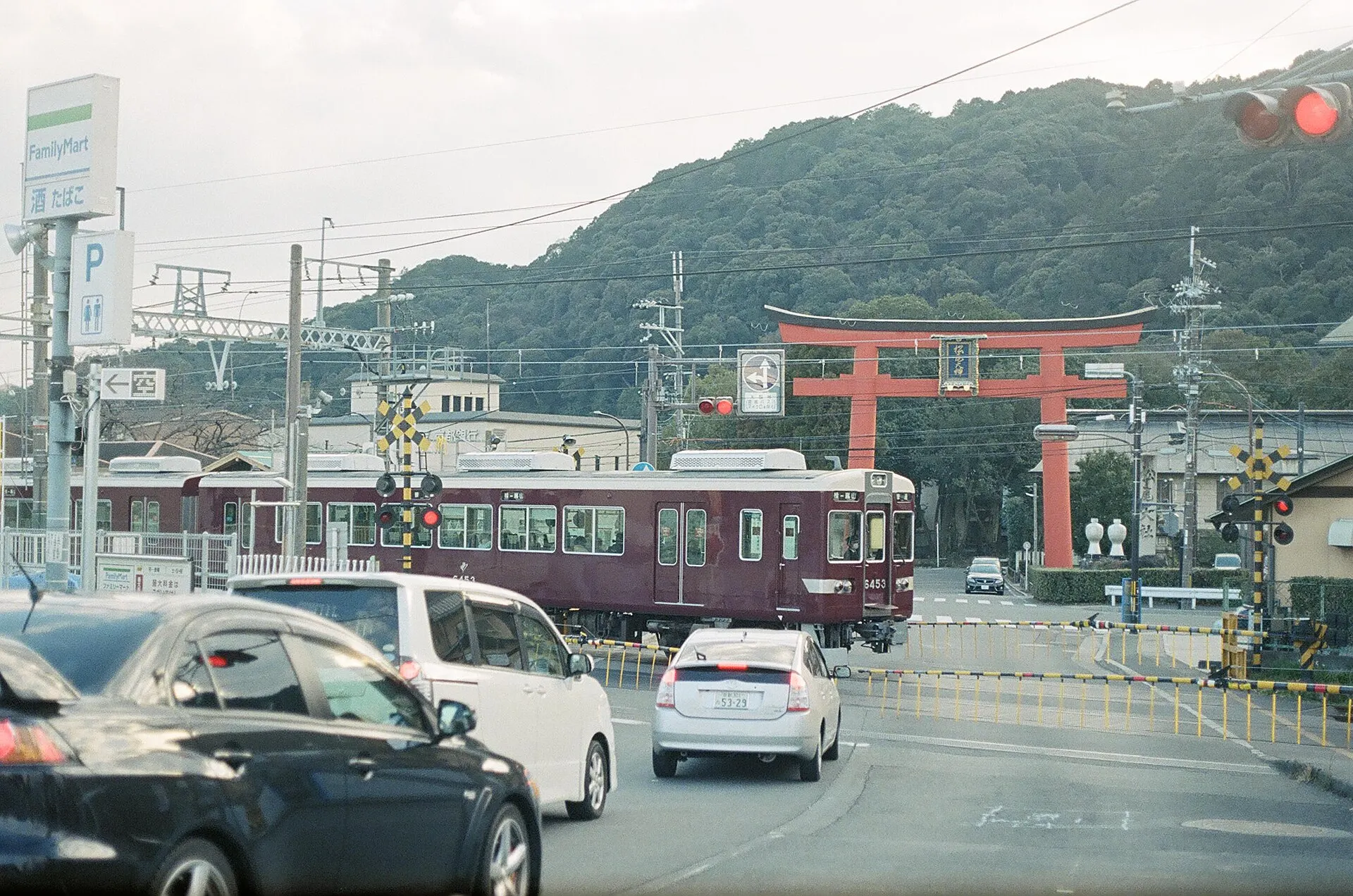 Red torii gate behind a road crossing with a maroon Hankyu train and parked cars