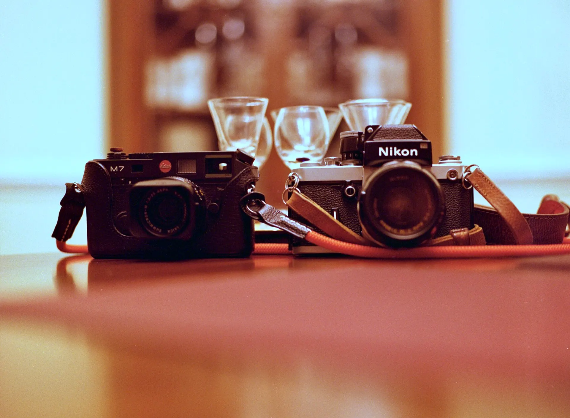 Two vintage film cameras on a wooden table with glasses blurred behind them