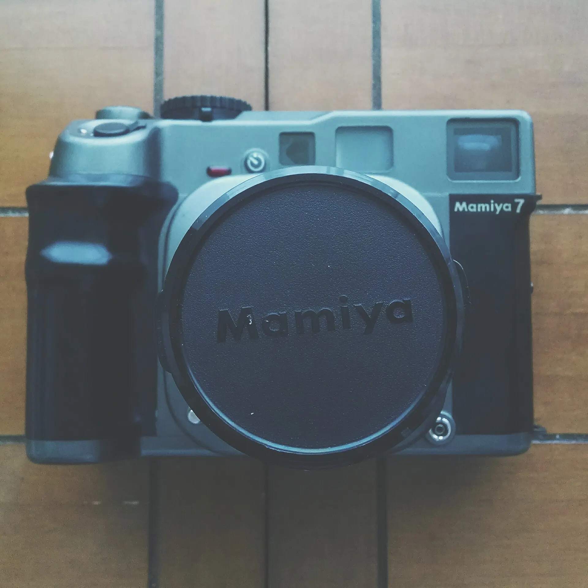 Mamiya 7 rangefinder seen from the front, with a Mamiya lens cap on the lens, lying on a wooden floor