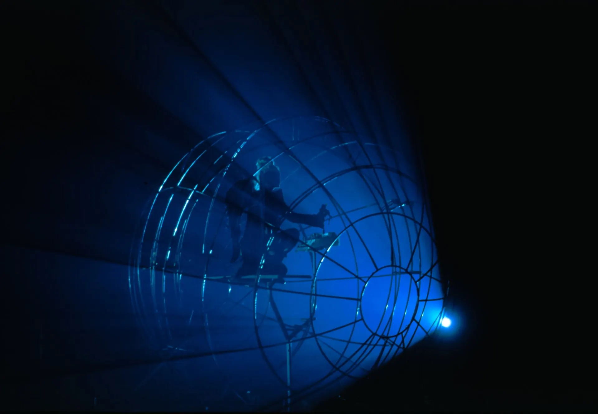 A street performer inside a large wheel lit by a blue spotlight at night