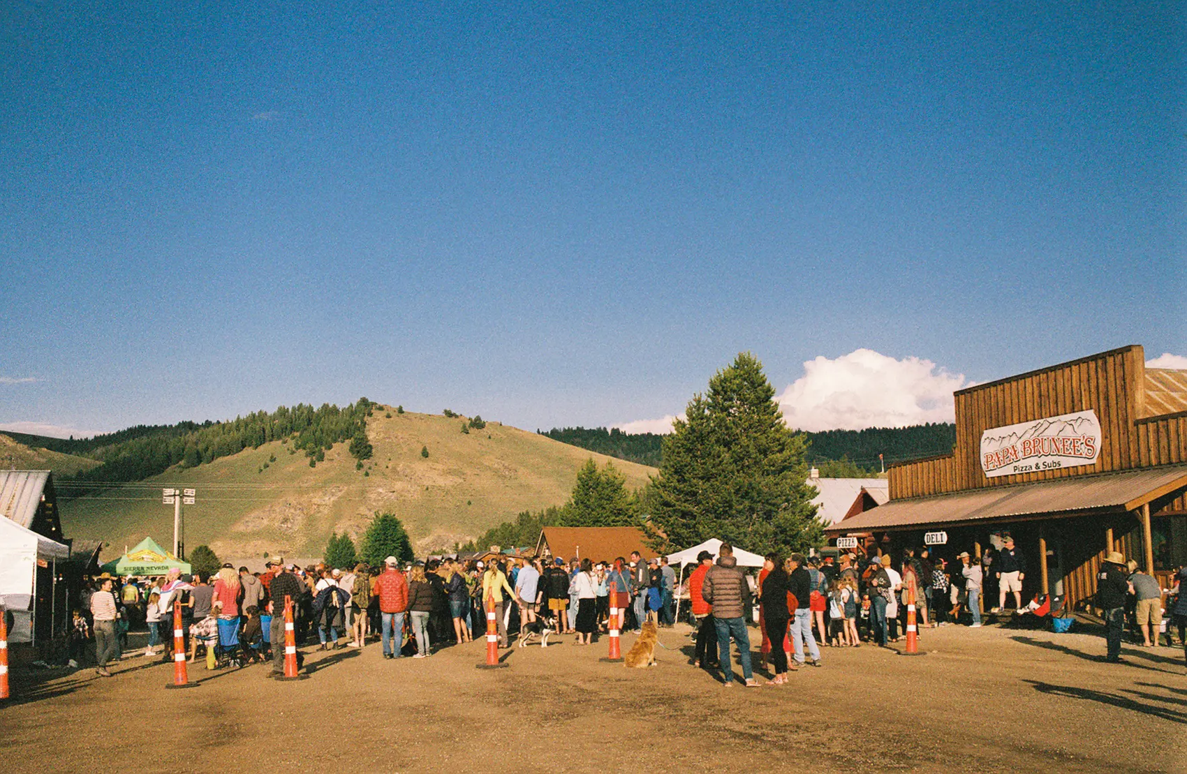 Crowd gathered on a dirt street in front of a wooden storefront, with green hills behind