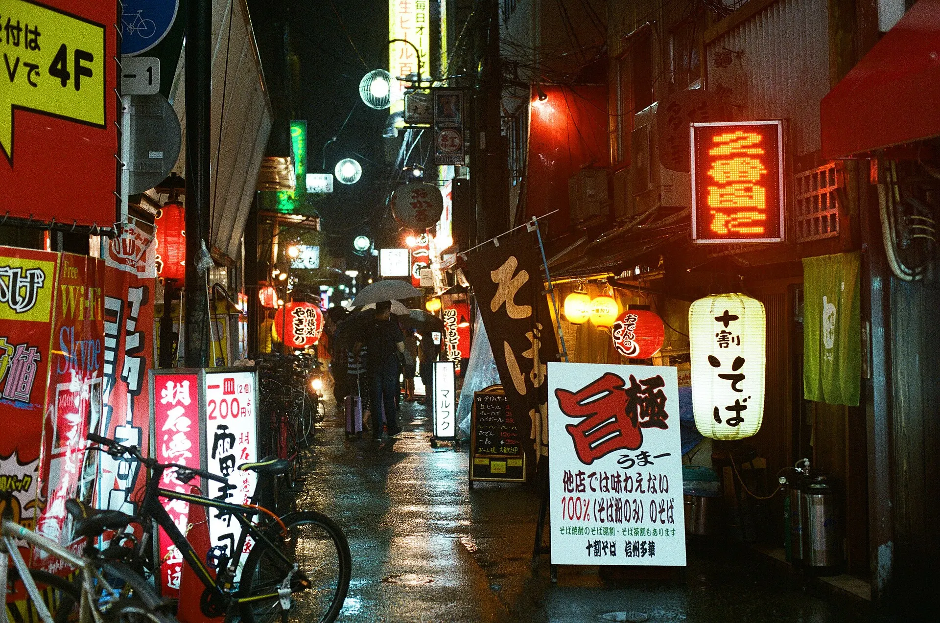 Narrow side street at night lit by red and yellow shop signs and paper lanterns