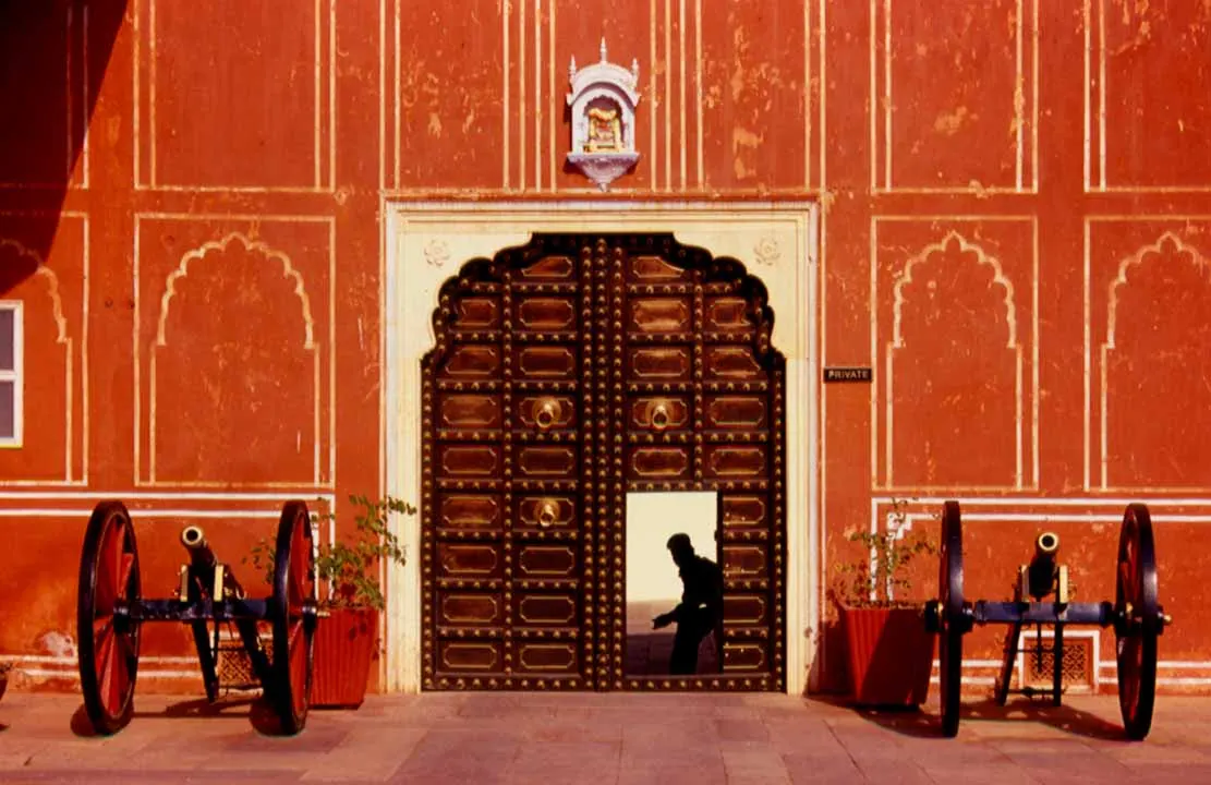 Brass-studded palace door in a terracotta wall, flanked by two cannons, with a figure silhouetted in the open doorway