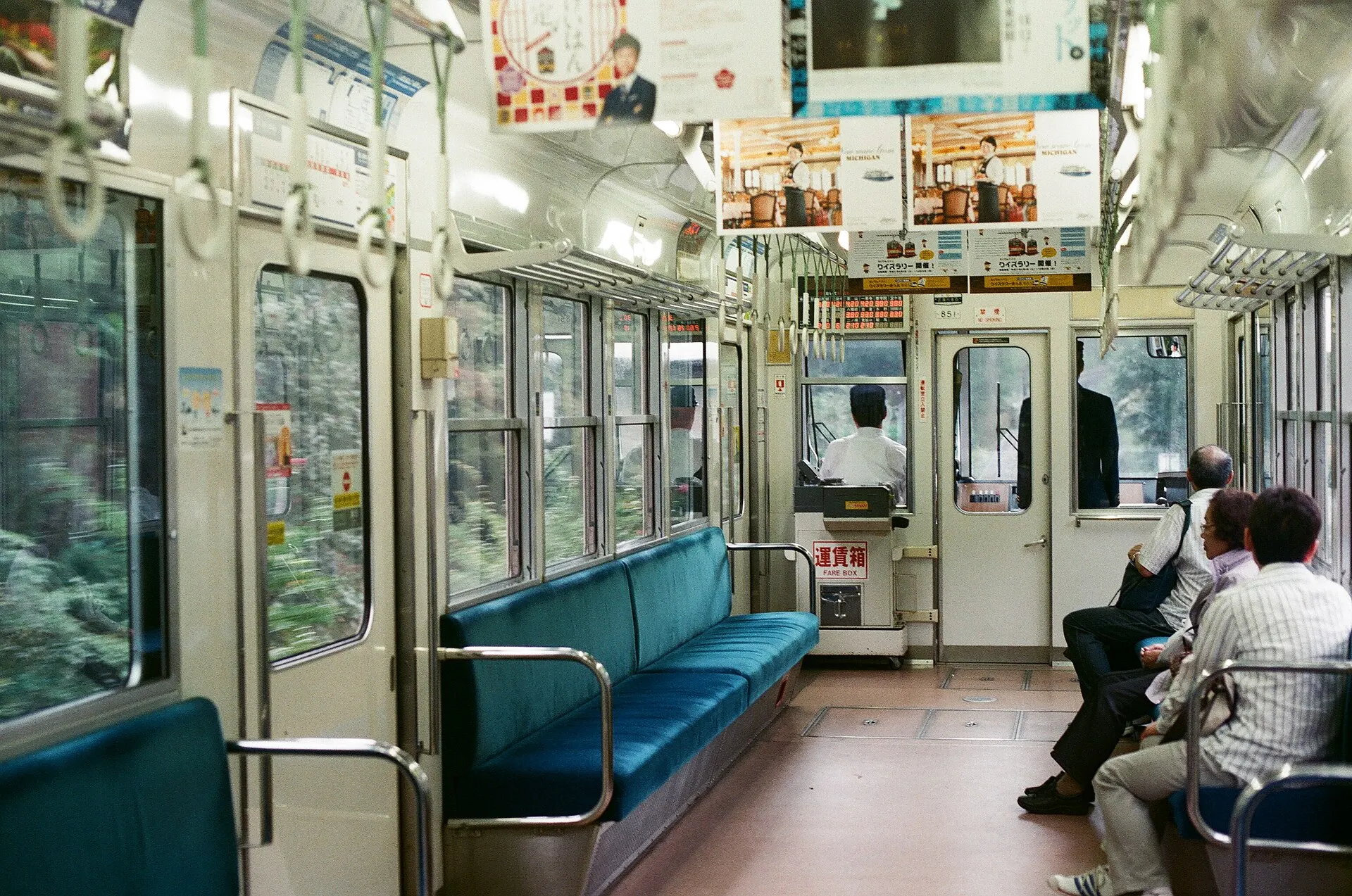 Inside a commuter train car with blue bench seats and a few seated passengers at the far end