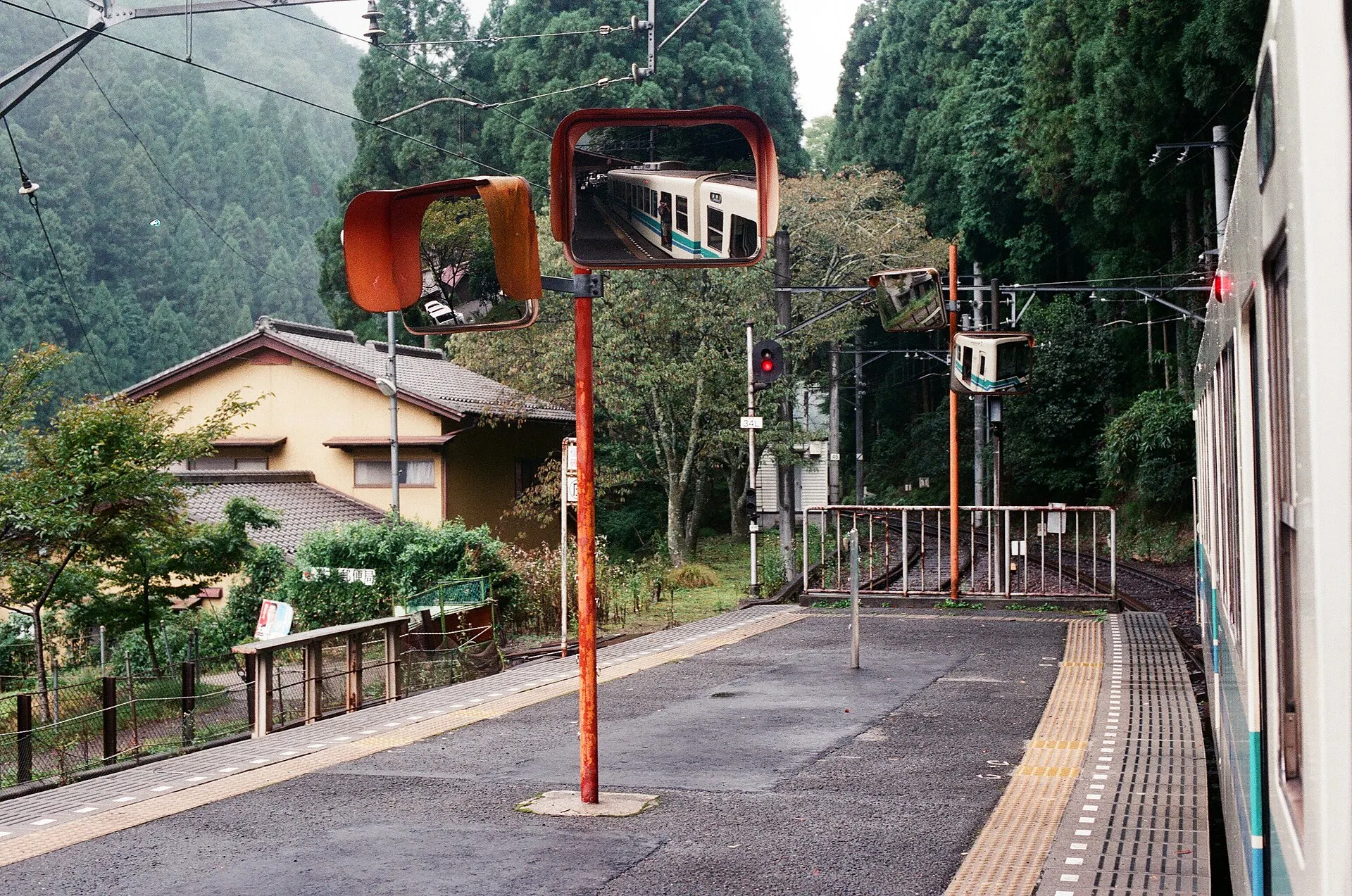 Empty station platform with a red mirror post, wooded hills and a house behind the fence