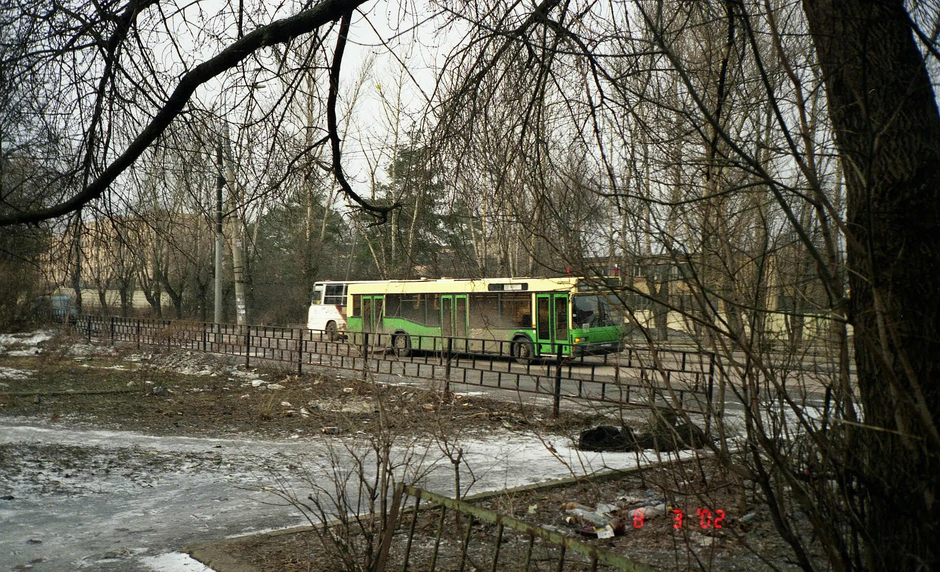 Green bus passing bare trees and a snowy riverbank